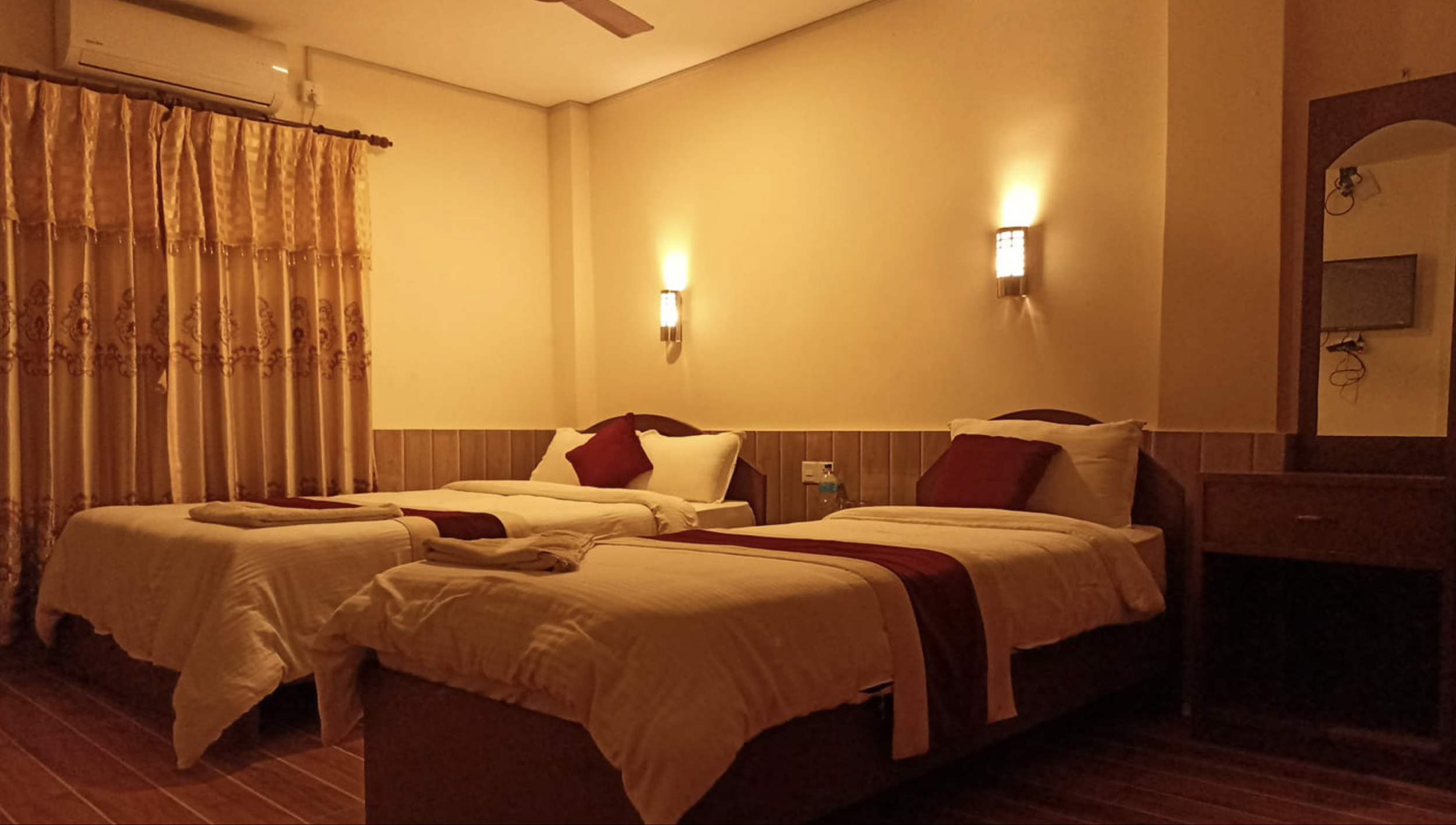 A dimly lit hotel room with two single beds, each with white linens and decorative red and white pillows. The beds are positioned side by side, with a small nightstand in between. The wall behind the beds has two mounted wall lights, and there is a window with ornate gold curtains on the left. A TV is visible in a mirror reflection on the right.
