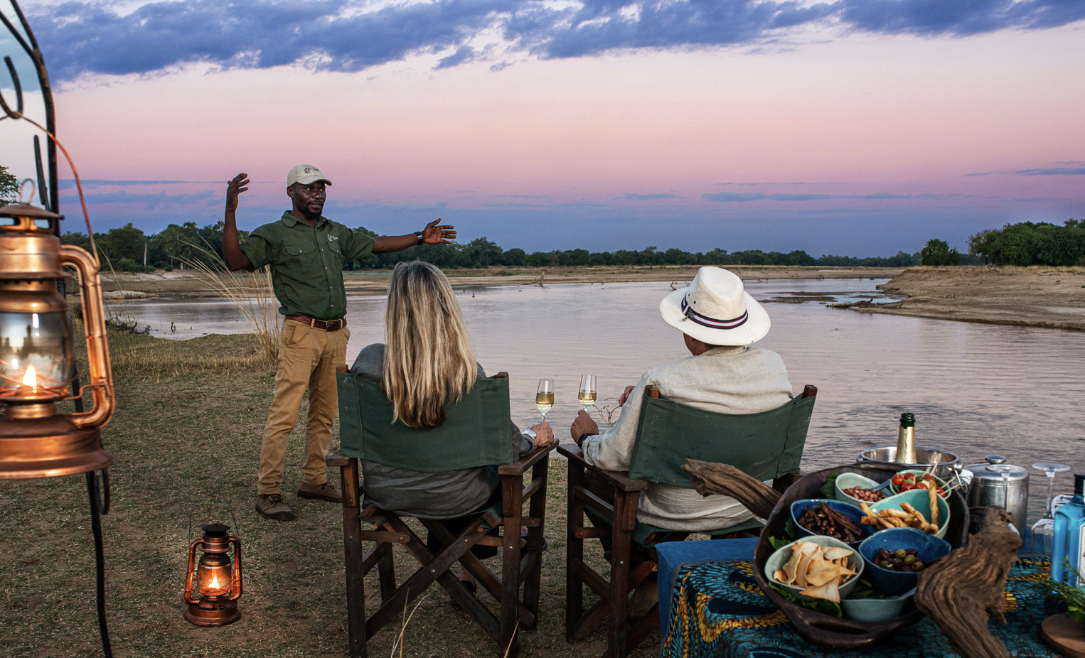 A man is giving a talk or presentation to two people sitting by a river at sunset. The scene includes drinks, snacks, and lanterns. Kaingo camp. Safari in South Luangwa national park with Wild Selection Tours.