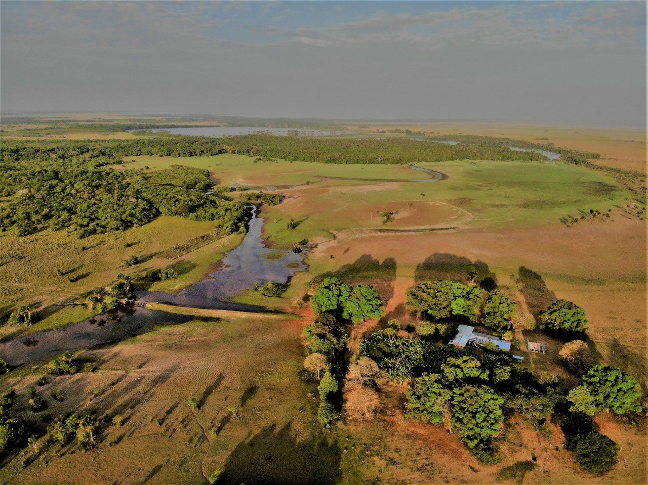 Aerial view of a rural landscape with water bodies, green fields, and trees, including a small building with a white roof.