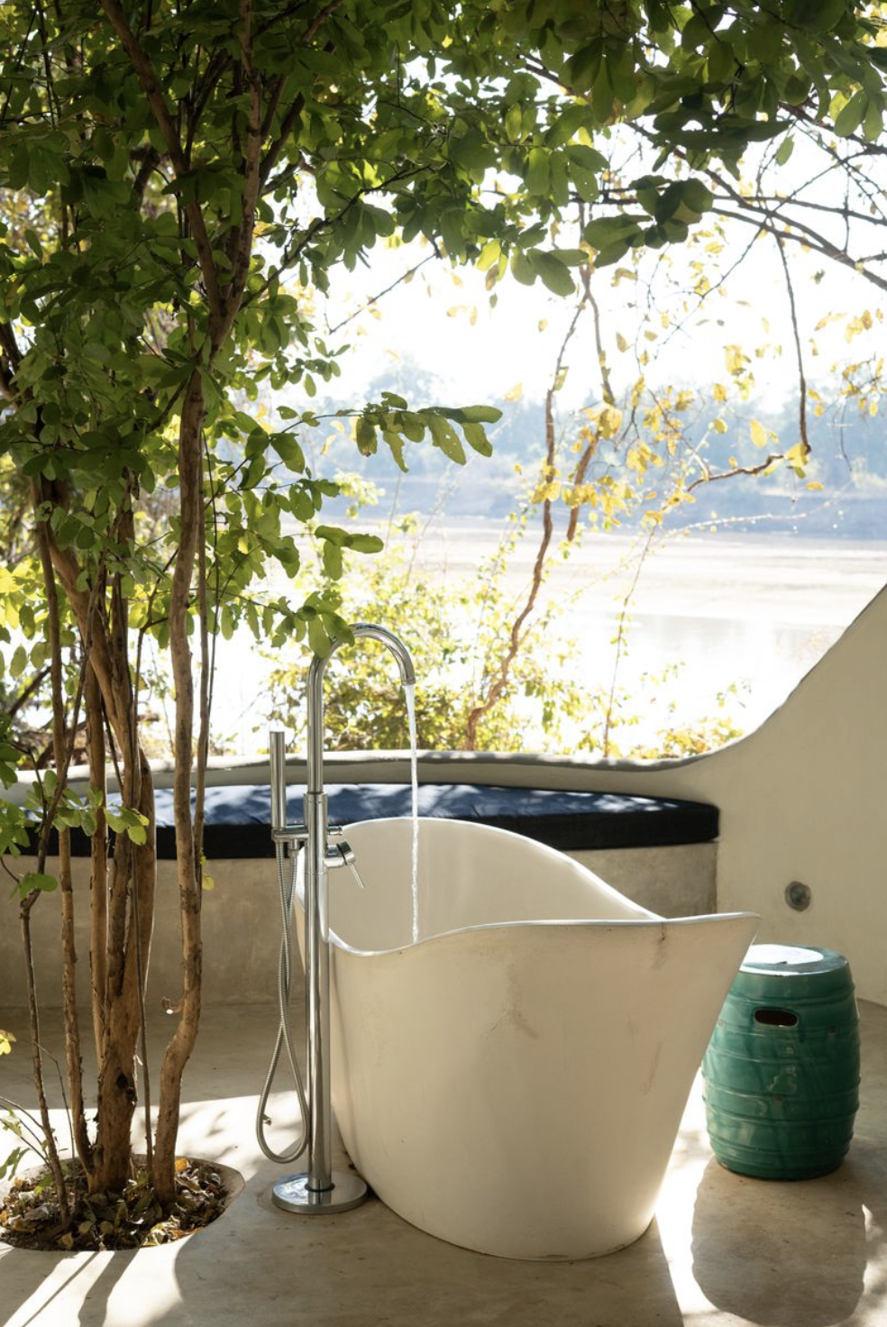 Outdoor bathtub on a patio overlooking a body of water, surrounded by green foliage and trees, with a green stool nearby. Kaingo camp. Safari in South Luangwa national park with Wild Selection Tours.