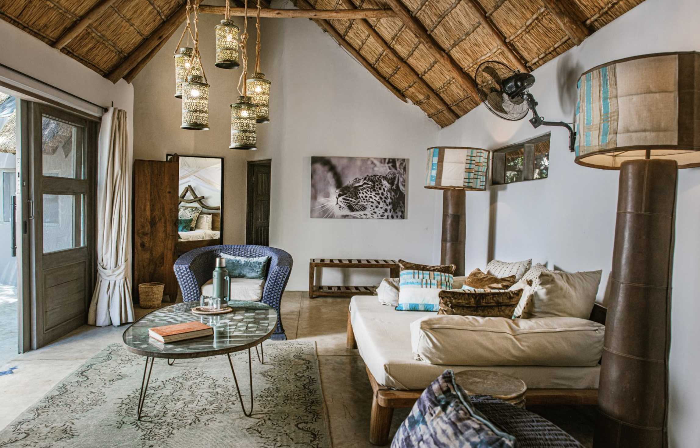 Living room with a high thatched roof, hanging lamps, a white sofa with various pillows, a round coffee table, a wicker chair, and a wall-mounted fan. There is a large black and white leopard photograph on the wall and a mirror next to a door with curtains.