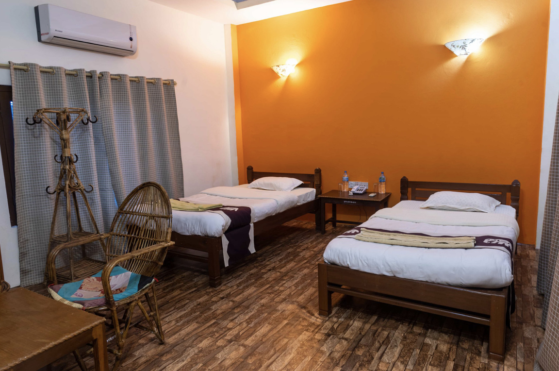 A hotel room with two single beds with white bedding, a bedside table with water bottles, a wooden chair with a cushion, a wicker coat rack, an air conditioner, and a window with checkered curtains. The room has a bright orange accent wall and wood flooring.
