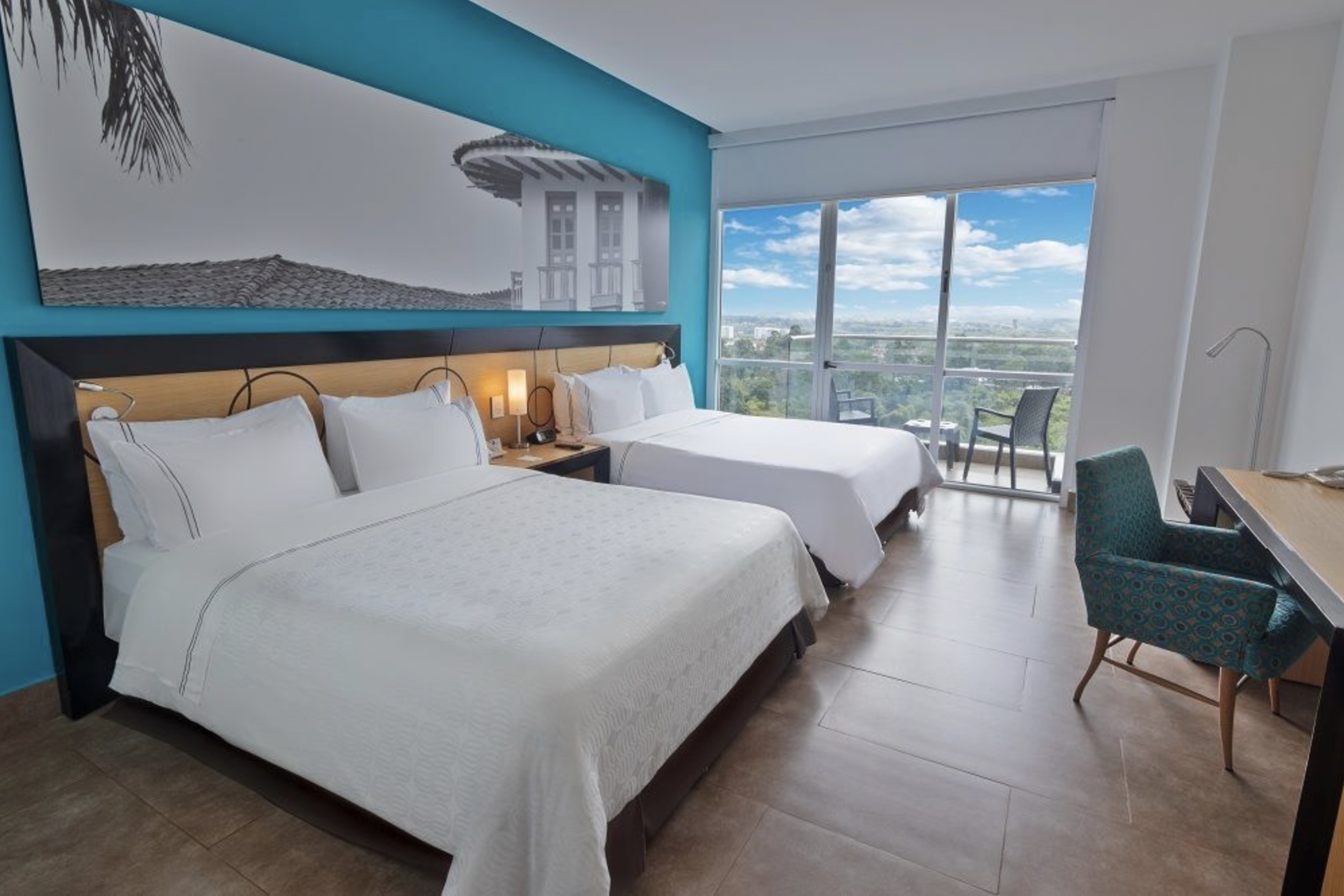 Hotel room with two beds, a balcony view of city and sky, blue accent wall, modern furniture, and natural light.