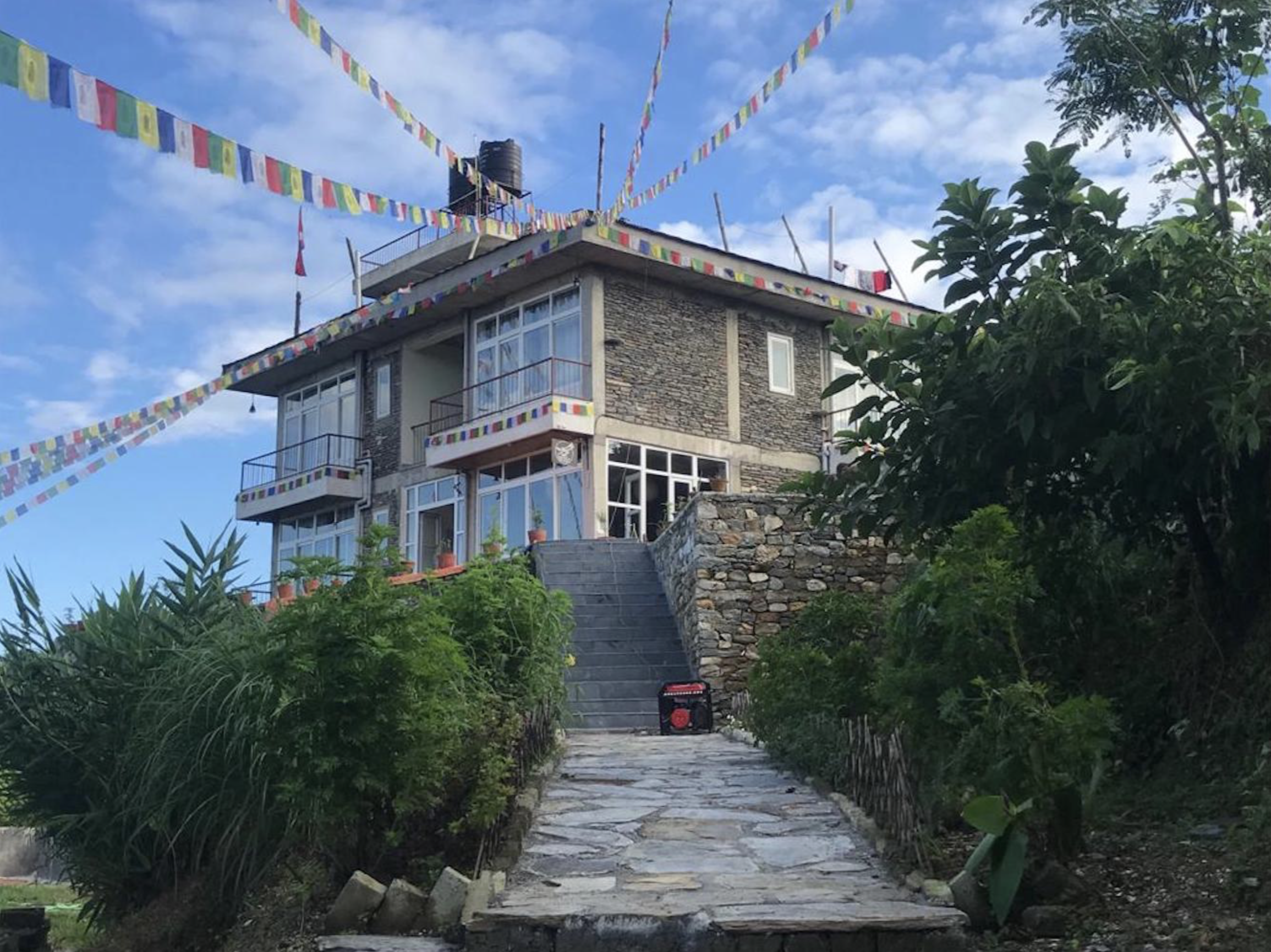 Stone house on a hill with colorful prayer flags hanging in the sky, surrounded by greenery and a stone pathway leading up to it, under a partly cloudy sky. Wildlife expedition and phot safari with Wild Selection Tours.