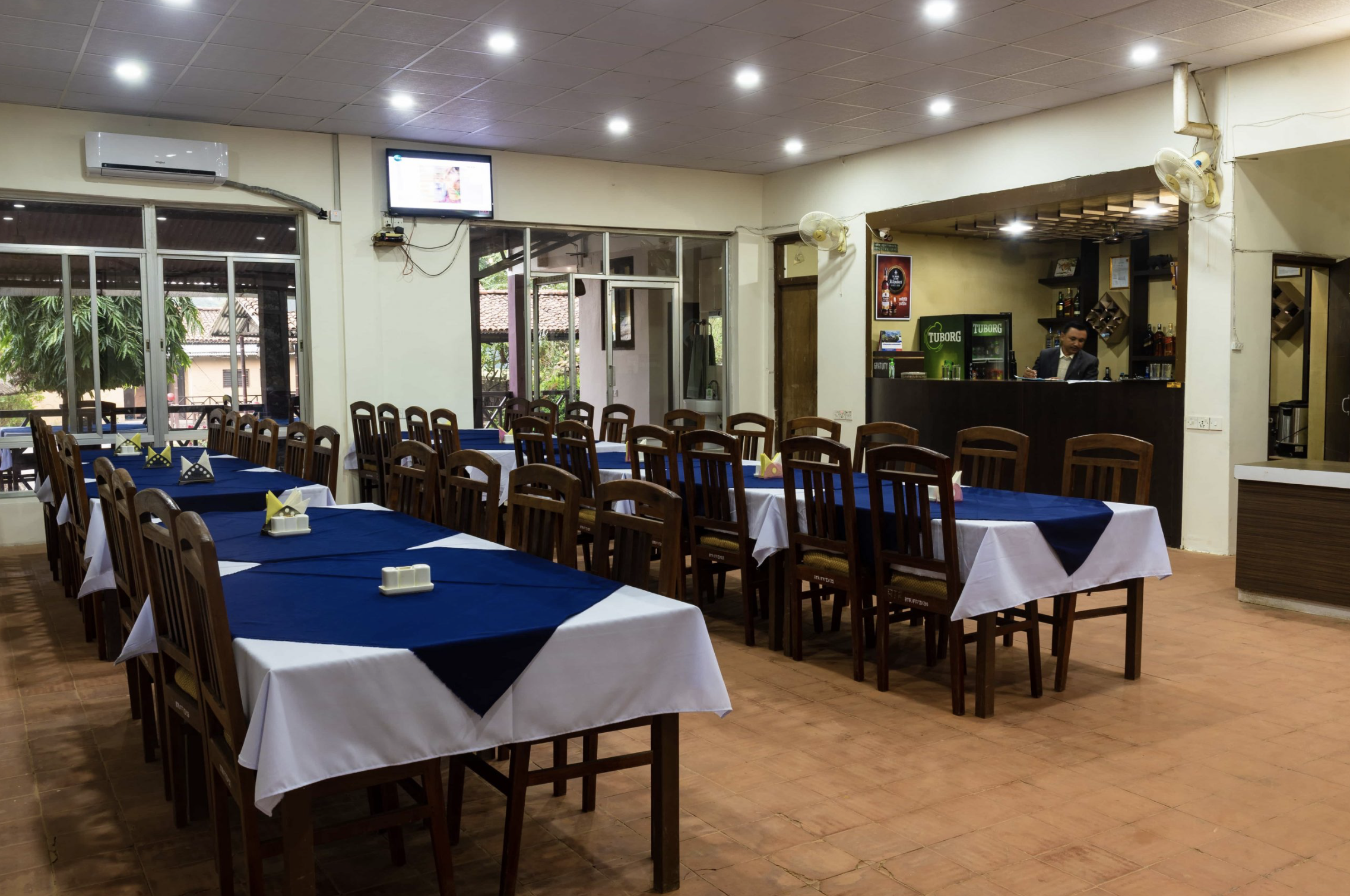 Restaurant interior with multiple tables covered with white and navy blue tablecloths, set for dining, with a bar in the background and large windows showing trees outside. Wildlife expedition and phot safari with Wild Selection Tours.