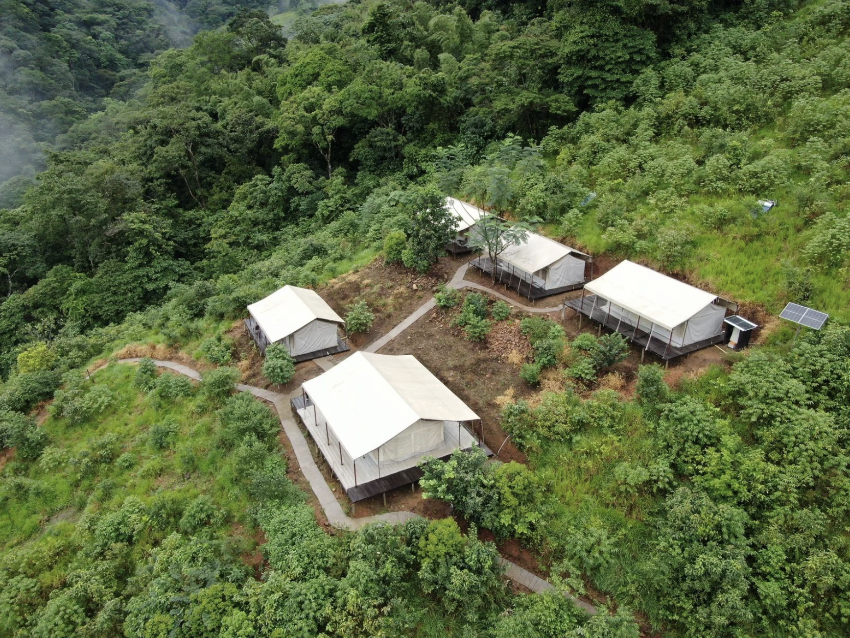 Aerial view of a small eco-resort with five white tents on stilts, situated on a lush green hillside surrounded by dense forest. A walking path connects the tents, and solar panels are visible near two of the tents.