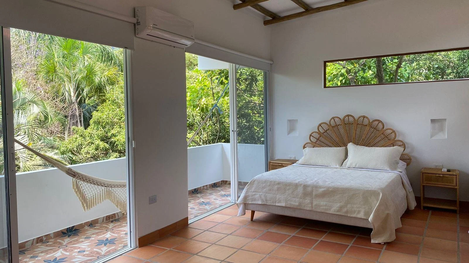 A bedroom with a white bed and pillows, wooden headboard, two nightstands, and sliding glass doors leading to a balcony with a hammock. The room has minimal decor and a window showing greenery outside.