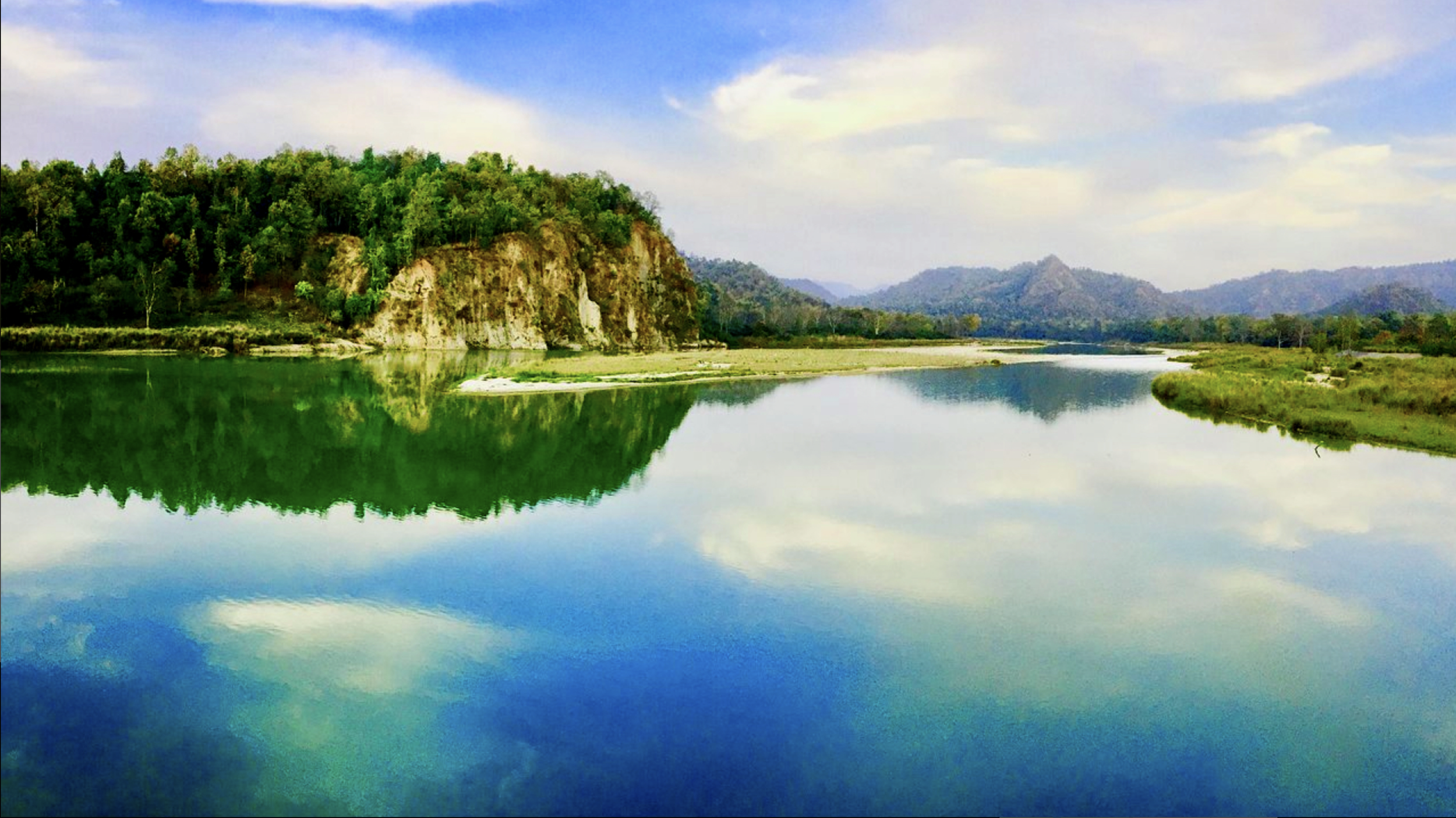 Calm river reflecting green trees and hills under a partly cloudy sky. Safari in Bardiya national park. Wildlife expedition and phot safari with Wild Selection Tours.