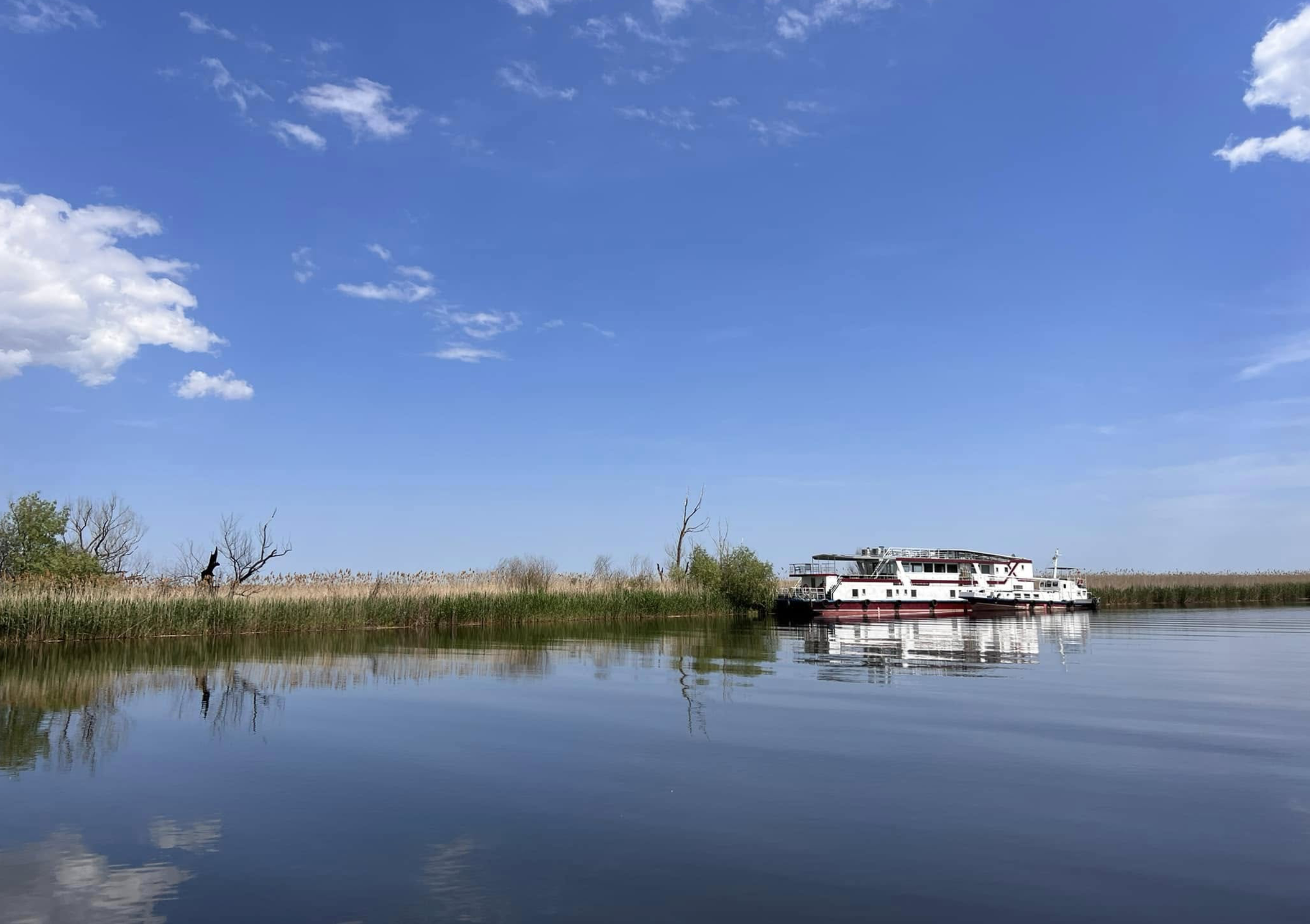 A clear blue sky with a few scattered white clouds over a calm river. On the riverbank, there are green trees and tall grasses. A white and red boat is docked on the right side of the river, reflected in the water.