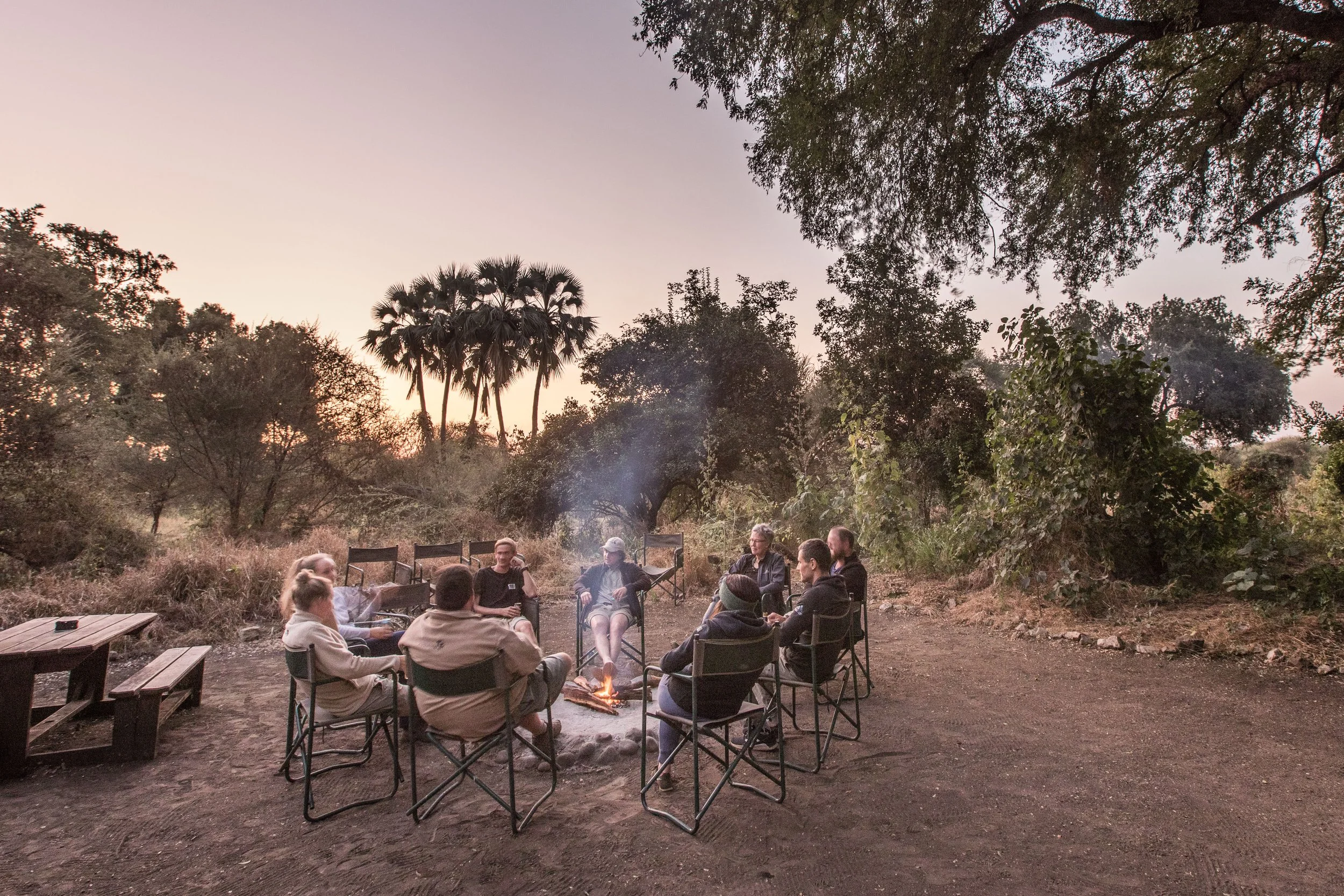 A group of people sitting around a campfire in a natural outdoor setting during dusk or early evening, surrounded by trees and vegetation.