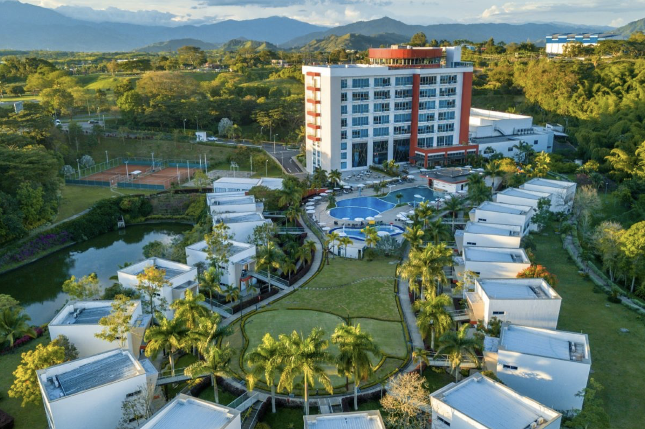 Aerial view of a resort or hotel complex with a large multi-story building, swimming pools, tennis courts, lush greenery, and surrounding mountains in the background.