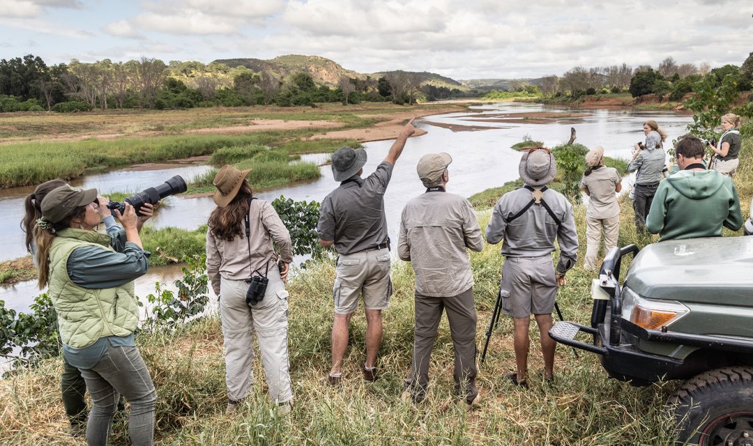 Group of people on a river bank, many with cameras or binoculars, observing a river and landscape.