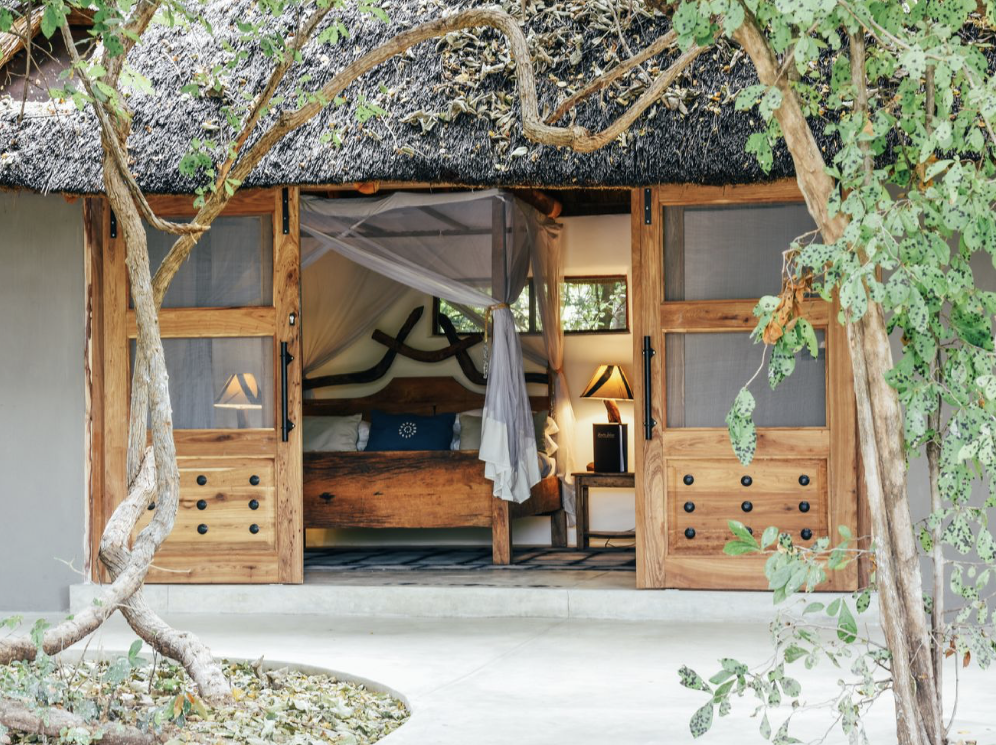 View of a rustic bedroom seen through open wooden doors, with a canopy bed, nightstand, lamp, and trees outside. Kaingo camp. Safari in South Luangwa national park with Wild Selection Tours.