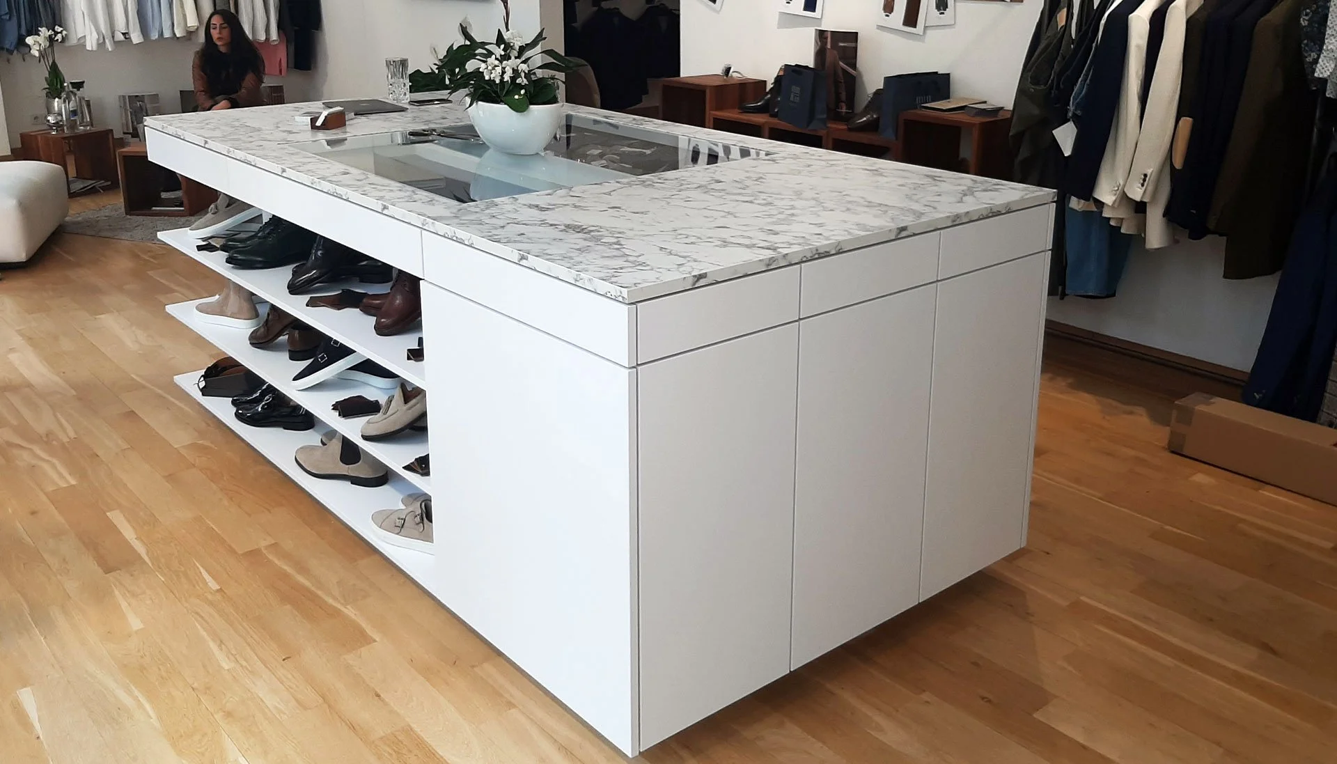 A modern store display featuring a white footwear island with drawers, a marble top with a flower vase, and shoes arranged on open shelves underneath. Clothing hangs in the background.