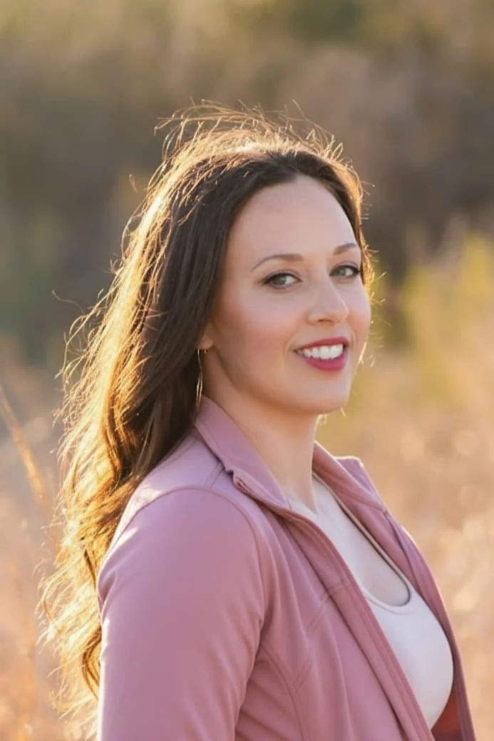 A young woman with long, wavy brown hair and fair skin smiling outdoors during sunset, wearing a light pink jacket and hoop earrings.