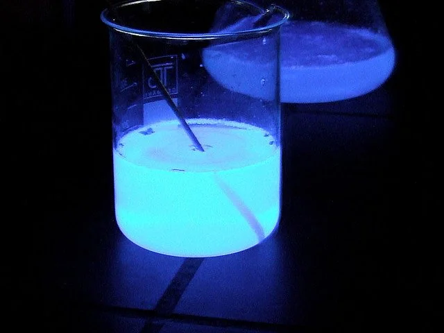 Un cylindre en verre contenant une substance fluorescente bleue, avec un agitateur en métal à l'intérieur, éclairé par une lumière UV dans l'obscurité.