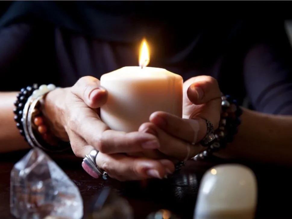 Person holding lit candle with both hands, wearing multiple bracelets and rings, with some crystals on the table in front.
