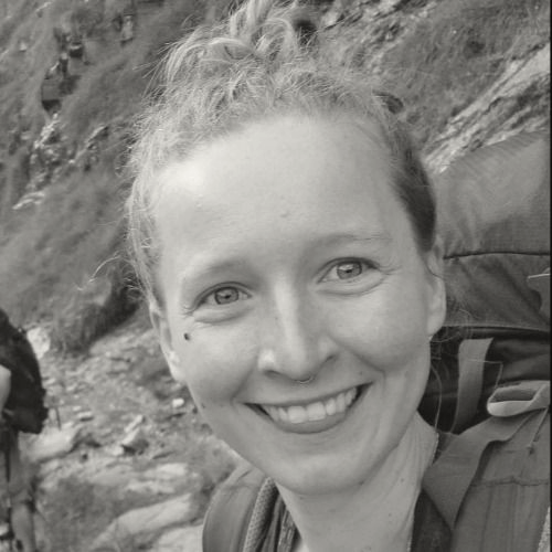 Portrait en noir et blanc d'une femme souriante avec un sac à dos, arrière-plan naturel de terrain rocheux et végétation.