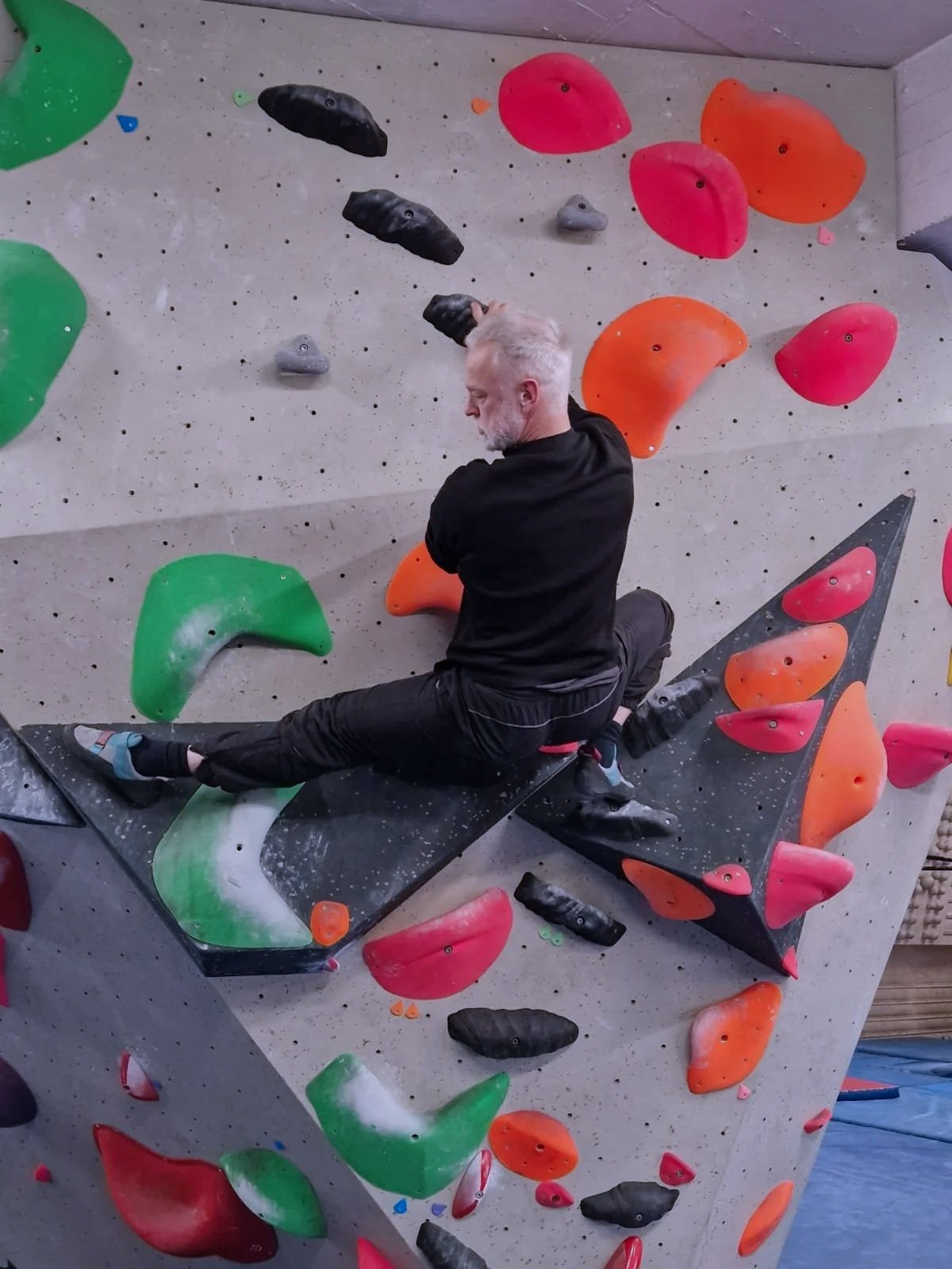 Homme à cheveux gris faisant de l'escalade sur un mur d'escalade intérieur avec des prises de différentes formes et couleurs, notamment rose, orange, vert et noir.