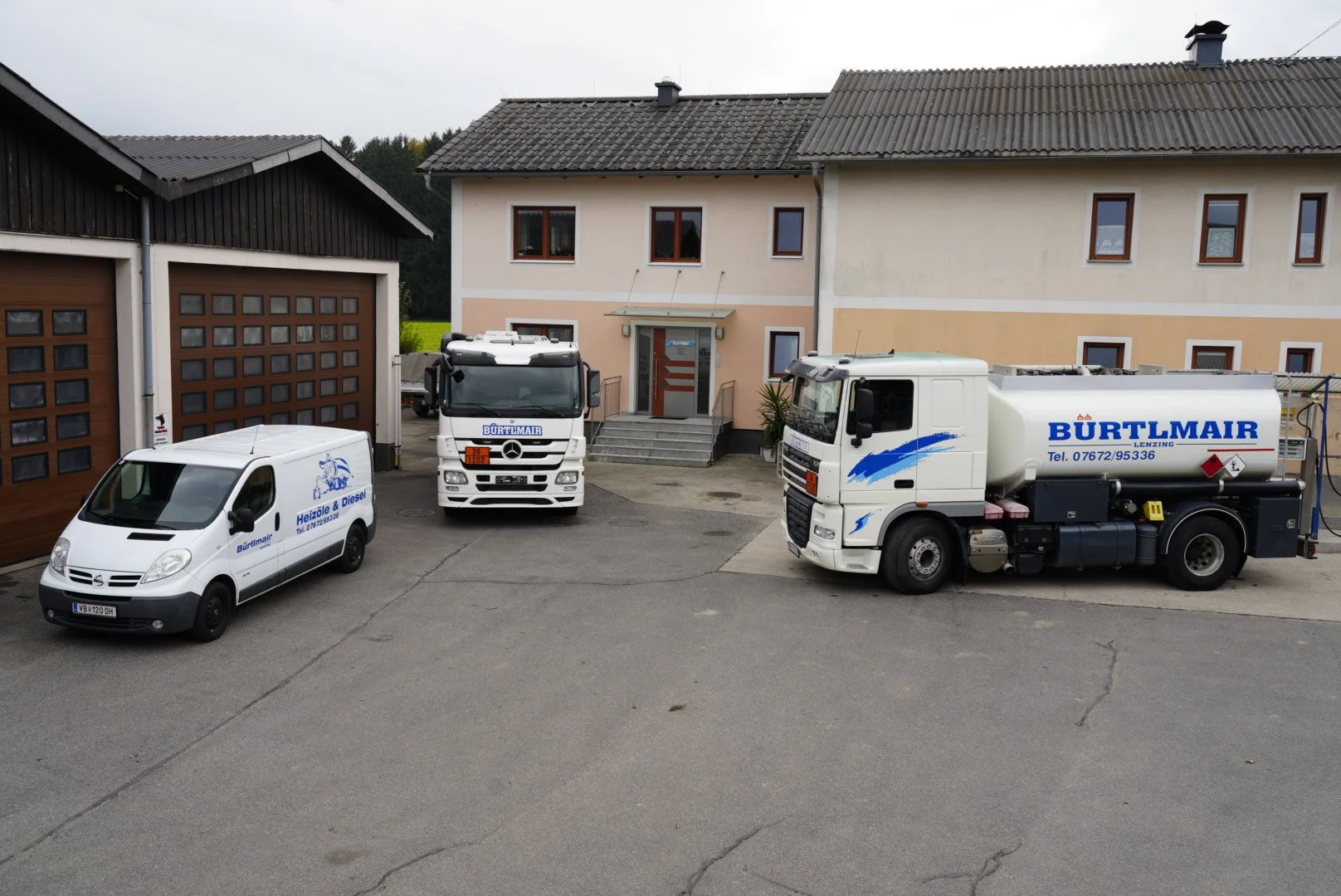 Zwei LKWs, eine weiße Firmenwagen, ein Haus mit zwei Etagen und eine Garage, davor stehen zwei Treppen, ein zweistöckiges Haus, die Fahrzeuge sind mit Firmenlogos 'Bürtlmair' und 'Helöl & Diesel' versehen.