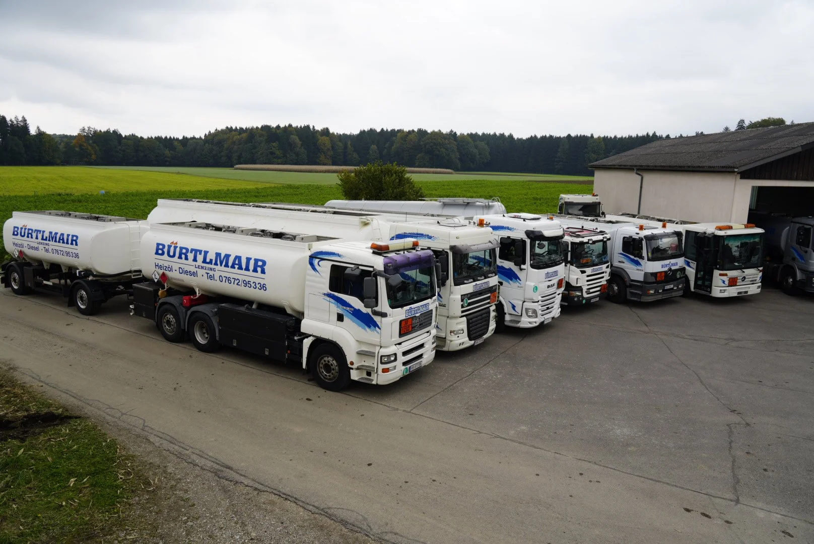 Mehrere weiße Tankwagen mit blauer Beschriftung parken auf einem Gelände, im Hintergrund grüne Wiesen und Bäume, über dem Himmel graue Wolken.