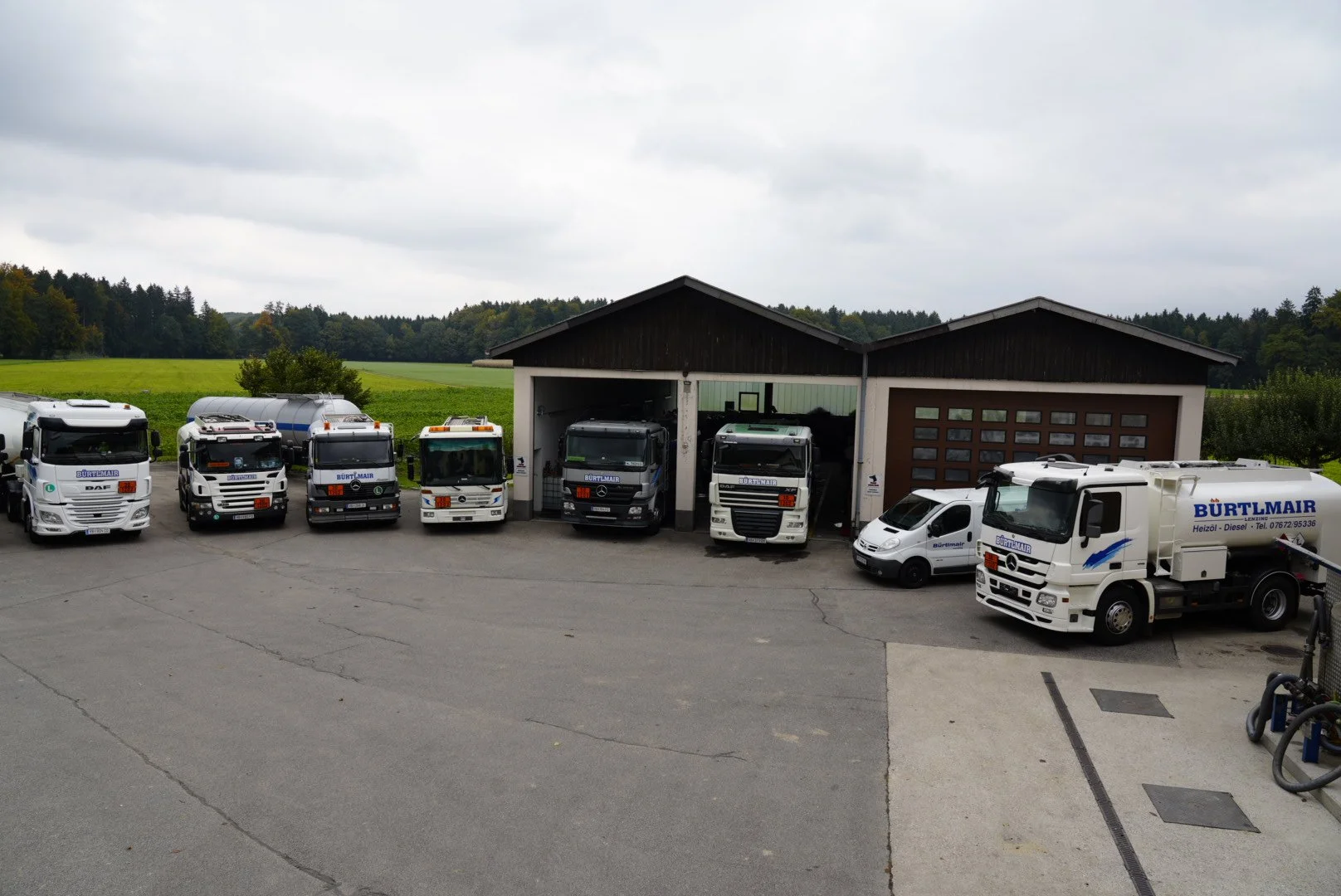 Lagerhalle mit mehreren LKWs und einem weißen Fahrzeug davor, auf einem Fahrplatz mit einer grünen Wiese im Hintergrund.