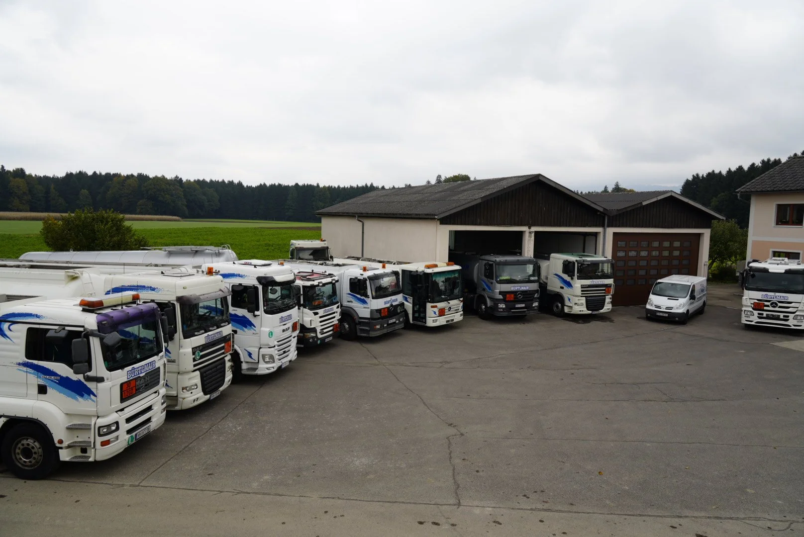 Mehrere weiße Transportfahrzeuge mit blauer Beschriftung sind auf einem Parkplatz vor einer Garage am Rande eines ländlichen Gebiets mit Wiesen und Bäumen zu sehen.