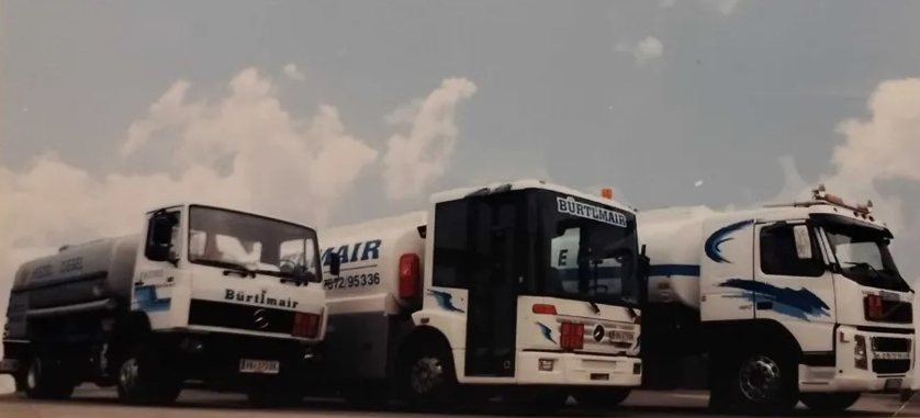 Drei weiße Fahrzeuge mit blauer Beschriftung und Streifen, darunter eine Tankstelle, stehen auf einer Straße bei bewölktem Himmel.
