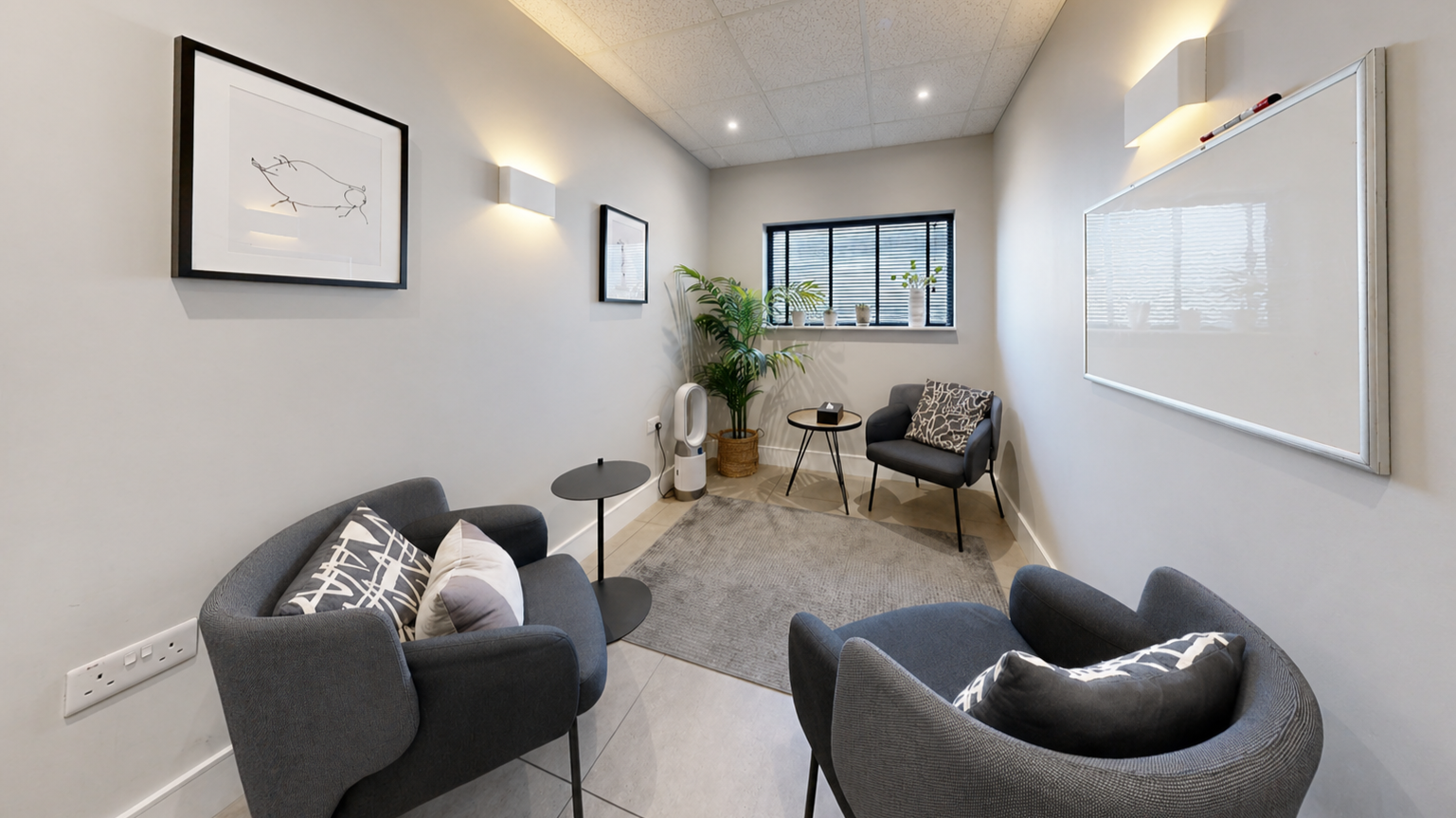 Small, modern office or waiting room with two gray chairs, a black armchair, a small round side table, a whiteboard on the wall, a window with blinds, potted plants, framed art, and a gray rug.