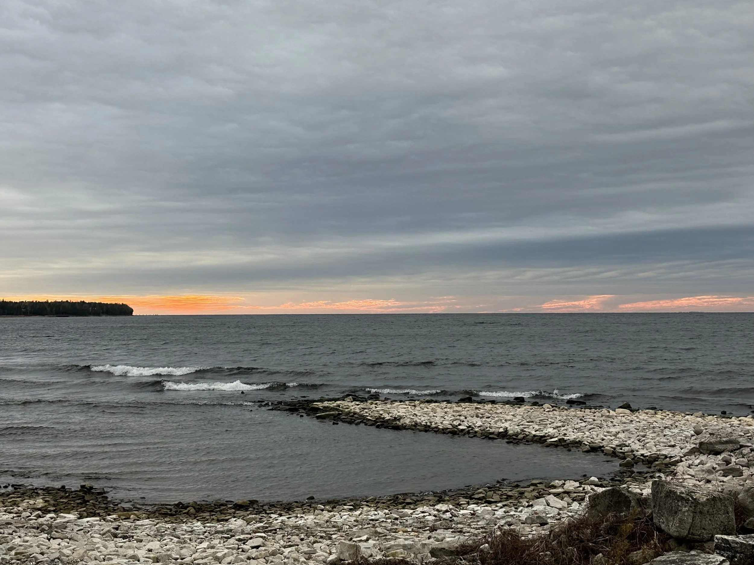 A rocky shoreline with small waves in the water, an overcast sky with a hint of sunset on the horizon, and a distant treeline to the left.