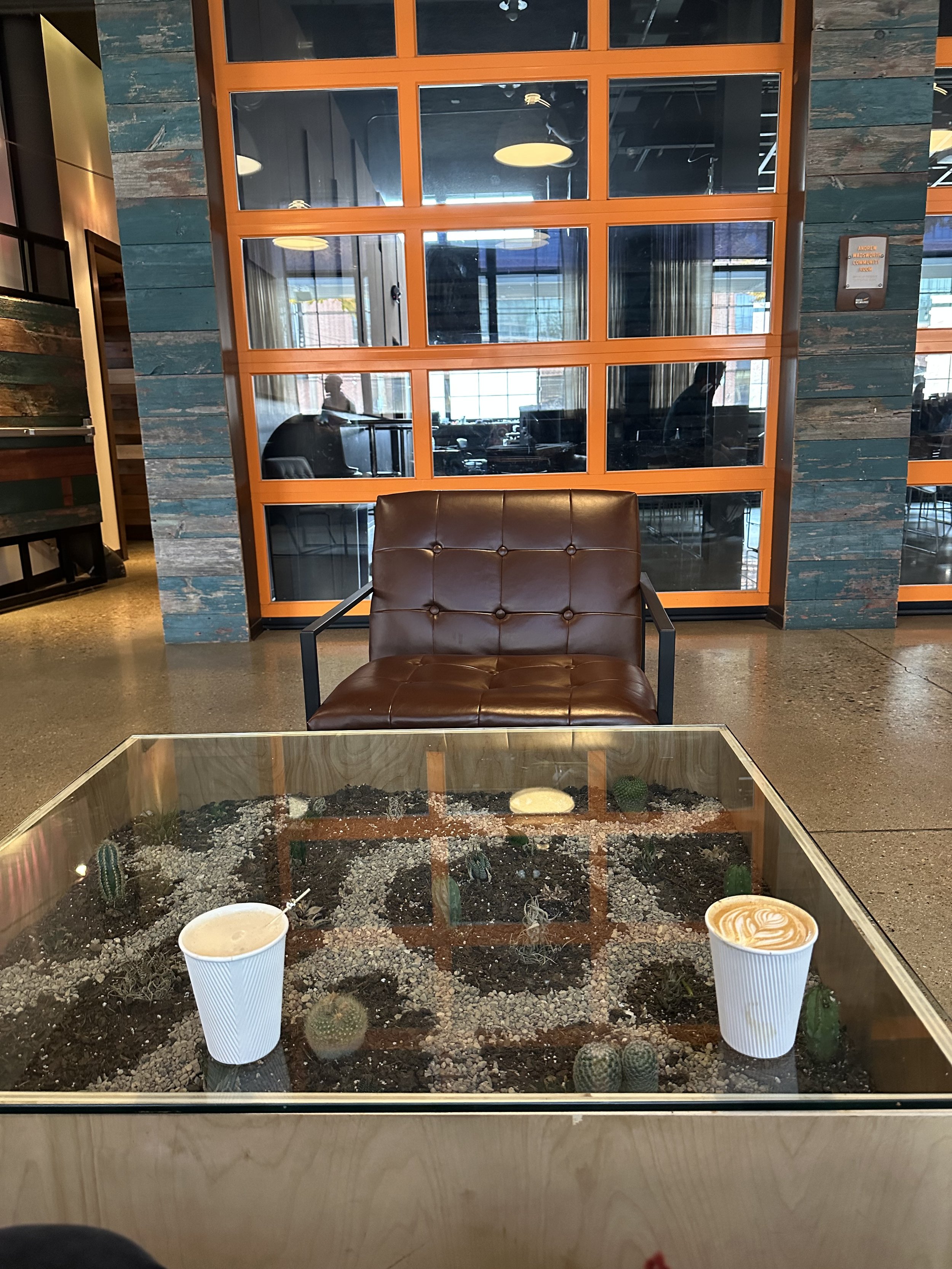 A cozy seating area with a leather armchair and a glass-top table filled with soil and small cacti, in front of a decorative wall with orange frames and a glass window showing a busy restaurant interior.