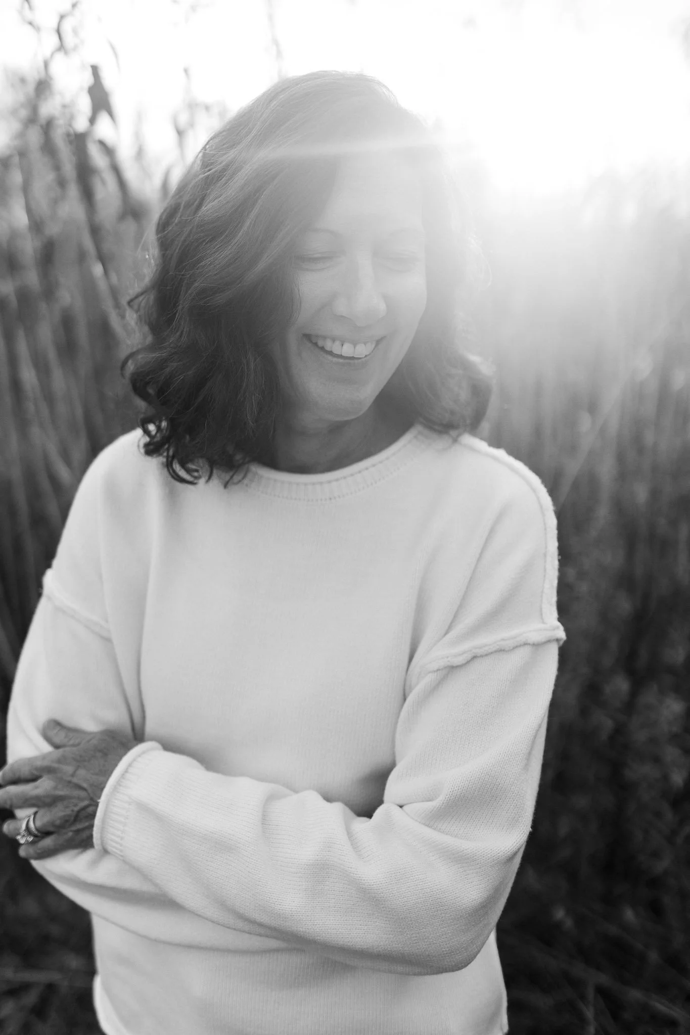 Black and white photo of Author and Speaker Debbie Prather smiling outdoors with her arms crossed, sunlight shining behind her.