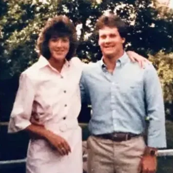 A woman and a man standing outside with trees in the background, smiling and posing together. The woman has dark curly hair and is wearing a white blouse, and the man has short hair and is wearing a light blue shirt.