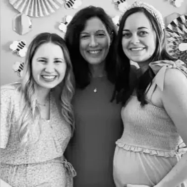 Three women smiling at a celebration with bee decorations in the background.