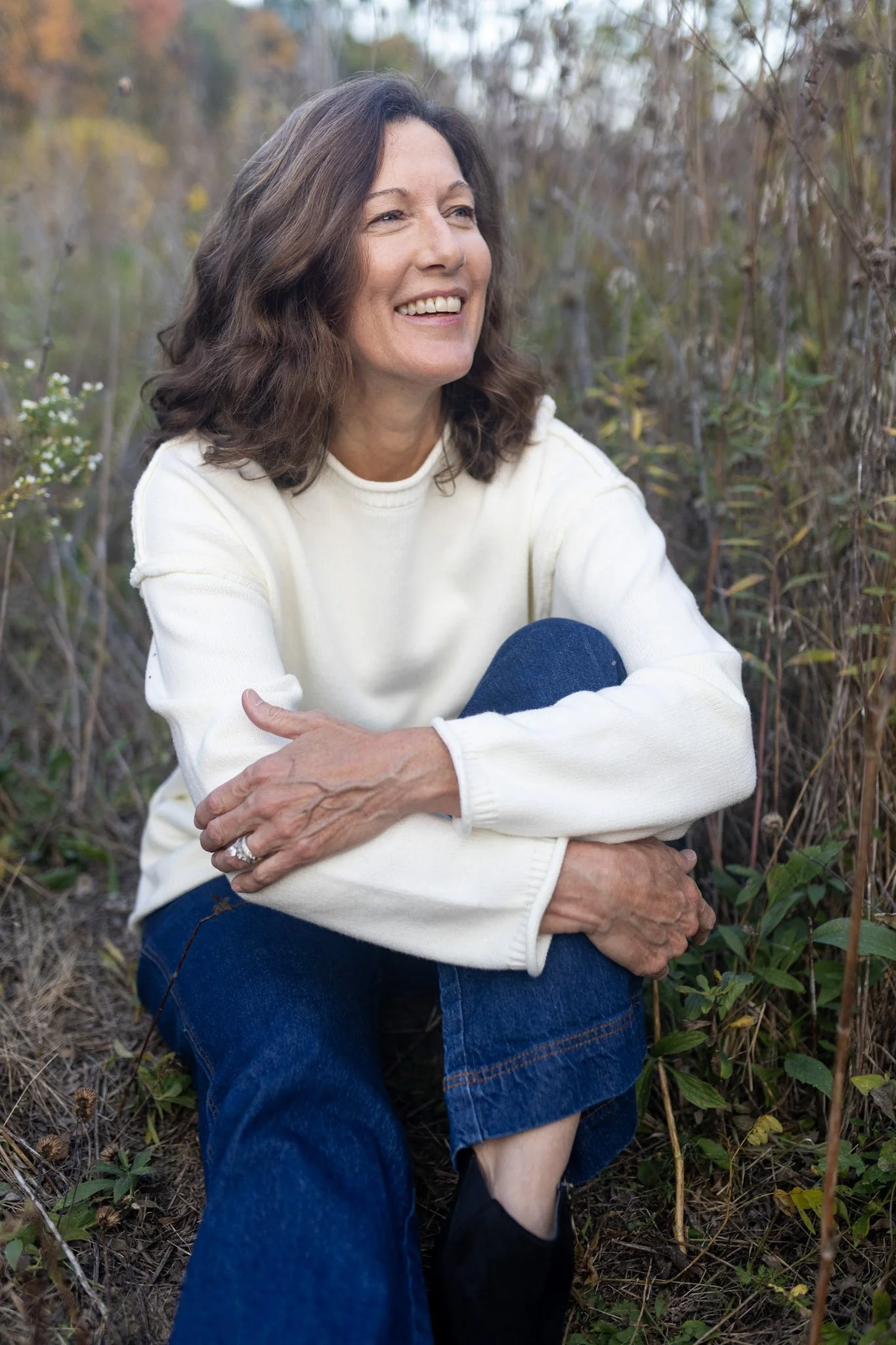 A woman with shoulder-length brown hair smiling outdoors, sitting among tall plants and trees, wearing a cream-colored sweater and blue jeans.