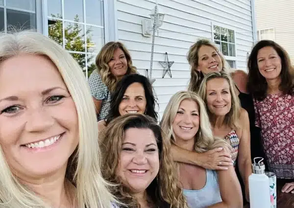 Group of women smiling and gathered outdoors in front of a house with white siding, with decorations on the wall.