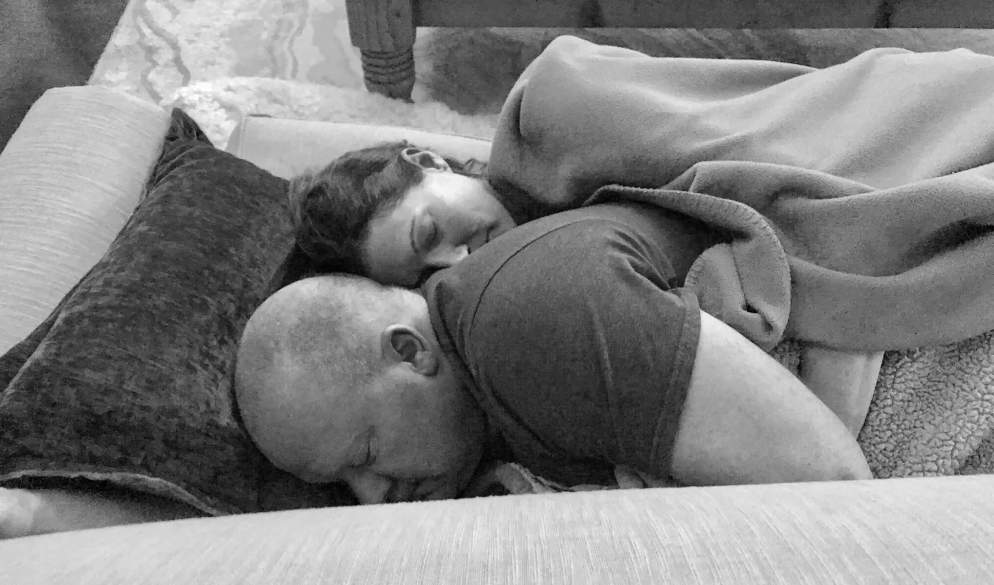 A black and white photo of a couple sleeping closely together on a couch, with a pillow and blanket.