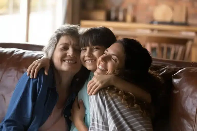 Three people, a woman, a girl, and a woman, smiling and hugging each other on a couch in a cozy living room. Debbieprather.com