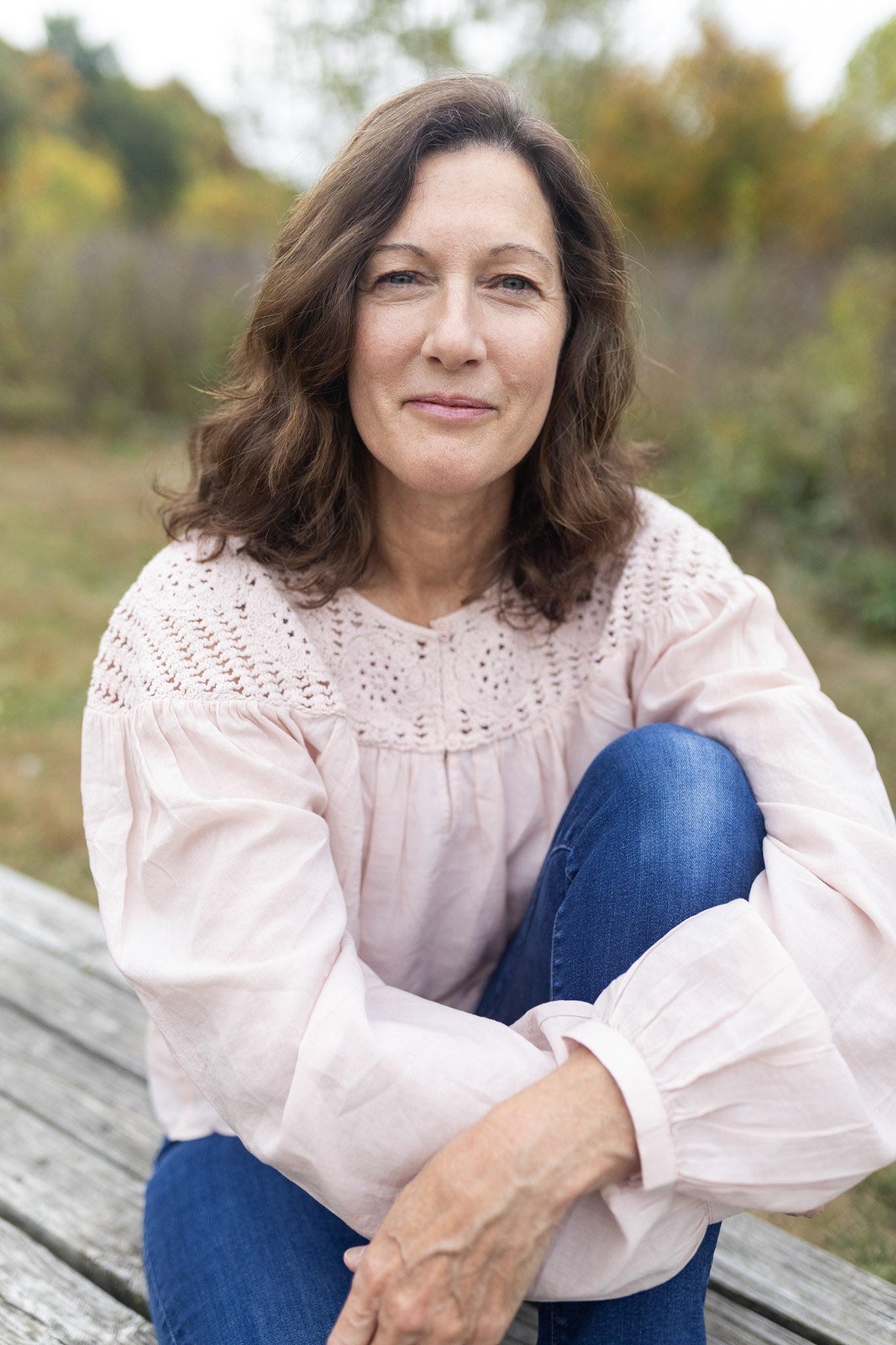 Author Debbie Prather with brown hair, wearing a light pink blouse and blue jeans, sitting on a wooden dock outdoors with fall foliage in the background.