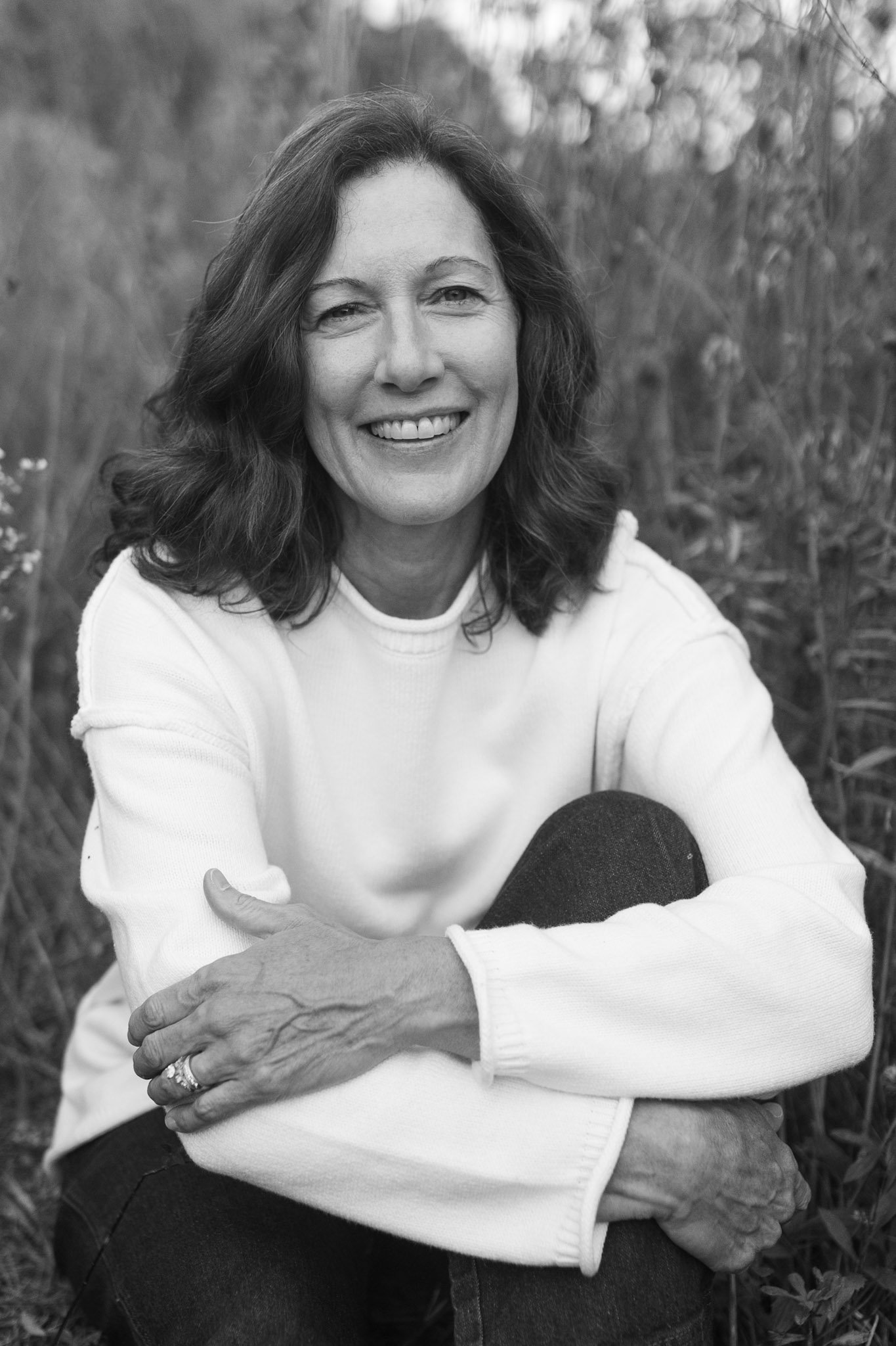 Black and white photo of a woman with shoulder-length wavy hair, smiling, sitting outdoors in a field. She is wearing a light-colored sweater and dark pants, with her arms crossed over her knees.