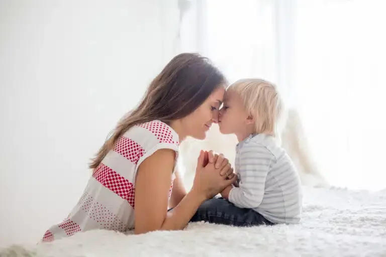 A woman and a young child with blonde hair touch foreheads and hold hands while sitting on a bed in a bright room with white walls and curtains.