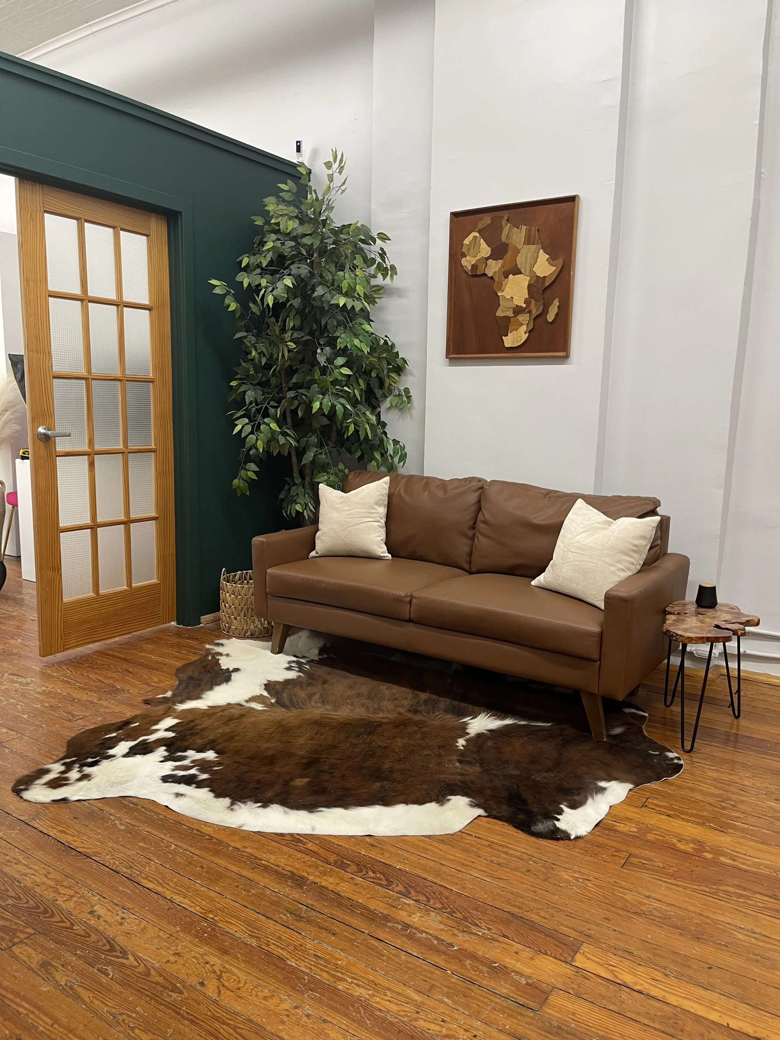 Cozy living room with brown leather sofa, white pillows, cowhide rug, large green plant, wooden framed map art, side table, and wooden door with glass panels.