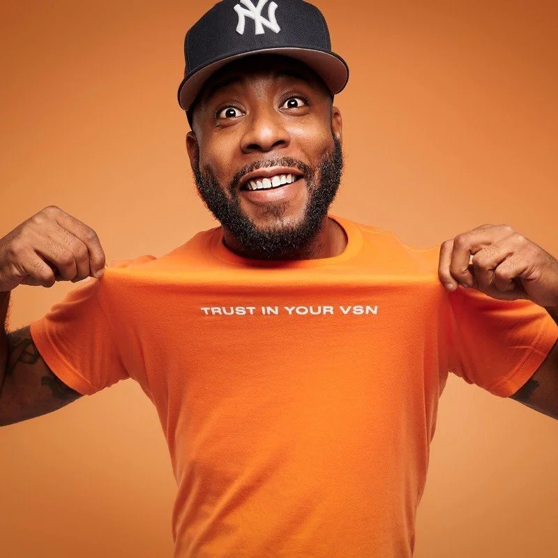 A man smiling and showing a bright expression, wearing a black baseball cap with a New York Yankees logo, an orange T-shirt with the text 'TRUST IN YOUR VSN', and lifting his T-shirt to reveal the message.
