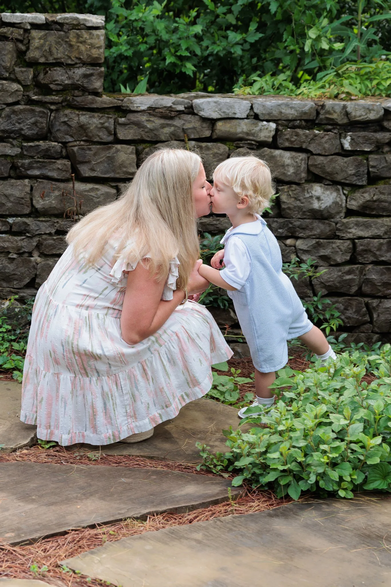 A woman and a young boy sharing a kiss outside near a stone wall and green bushes.