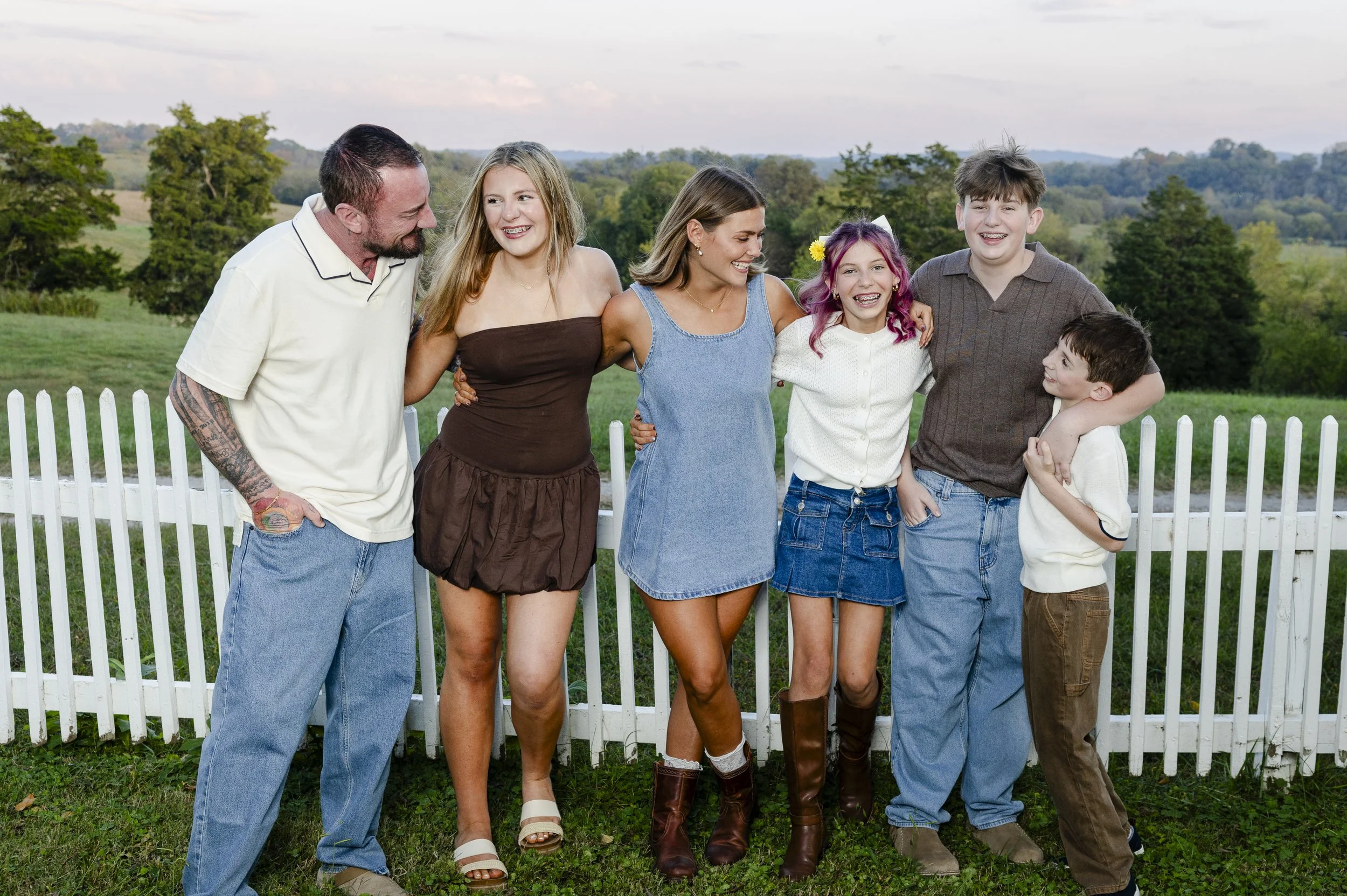 A family of six standing behind a white picket fence in a rural outdoor setting, smiling and interacting, with trees and hills in the background.