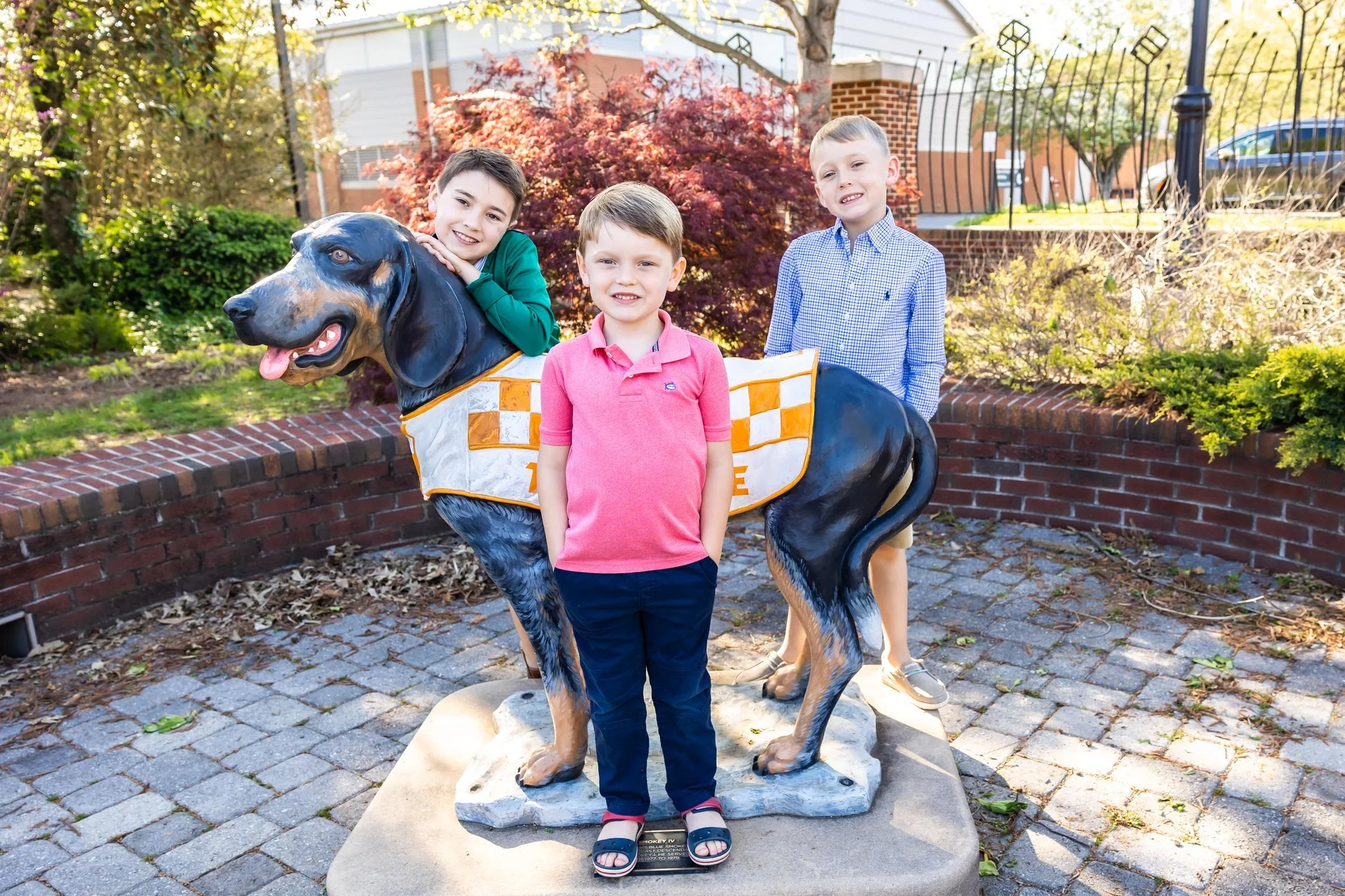 Three children standing next to a large statue of a black and tan dog, with a young girl in a pink polo shirt in front, smiling, and a boy in a green shirt and two boys in blue shirts behind, in an outdoor park setting with trees and a brick and metal fence in the background.