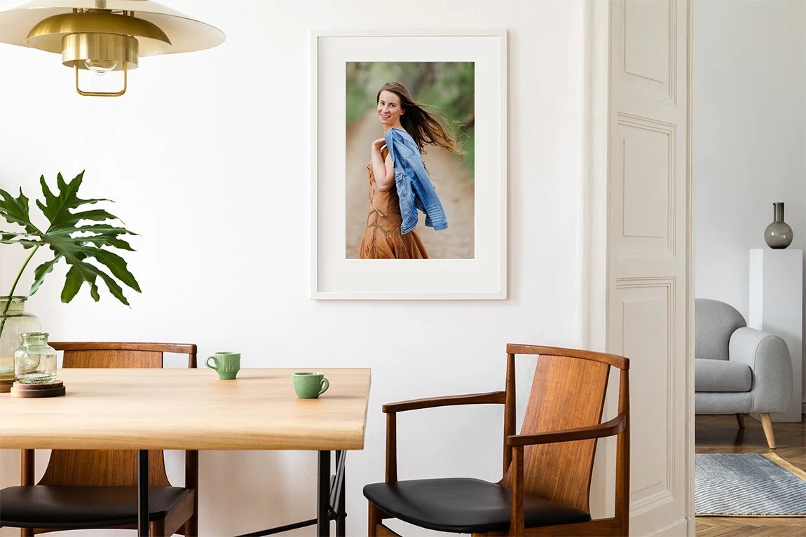 A dining room with a wooden table and chairs, a large framed portrait of a woman on the wall, green mugs on the table, green plant in a glass vase, and a gold ceiling light fixture.