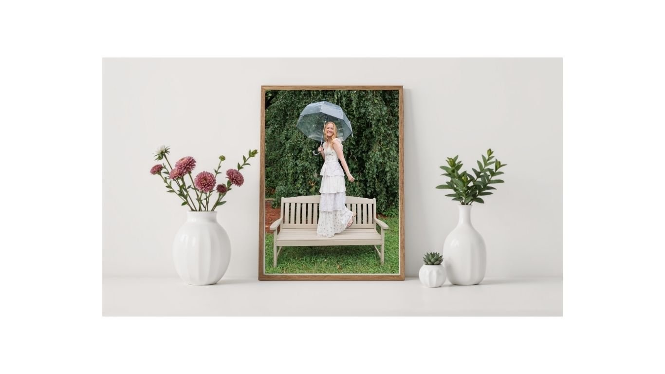 A framed photo of a smiling woman in a white dress holding an umbrella, standing on a bench outdoors with green trees in the background. The framed photo is surrounded by white vases with flowers and greenery on a white surface.