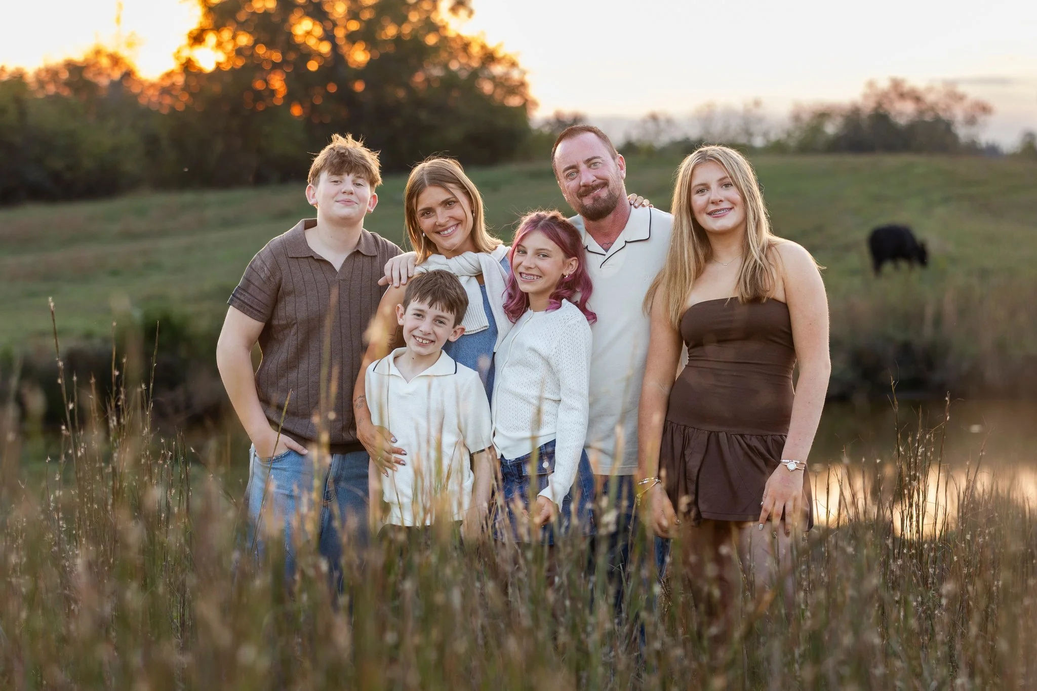 Family of six standing outdoors during sunset, smiling at the camera. Trees in the background, tall grass in the foreground.