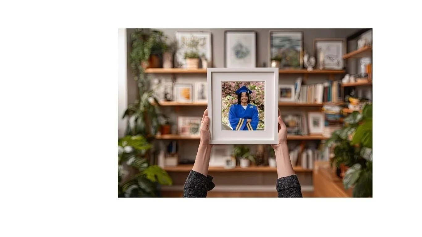 Person holding a framed photo of a smiling woman in a blue graduation gown and cap, with blossoming trees in the background, in a cozy, decorated living room with shelves filled with books and plants.